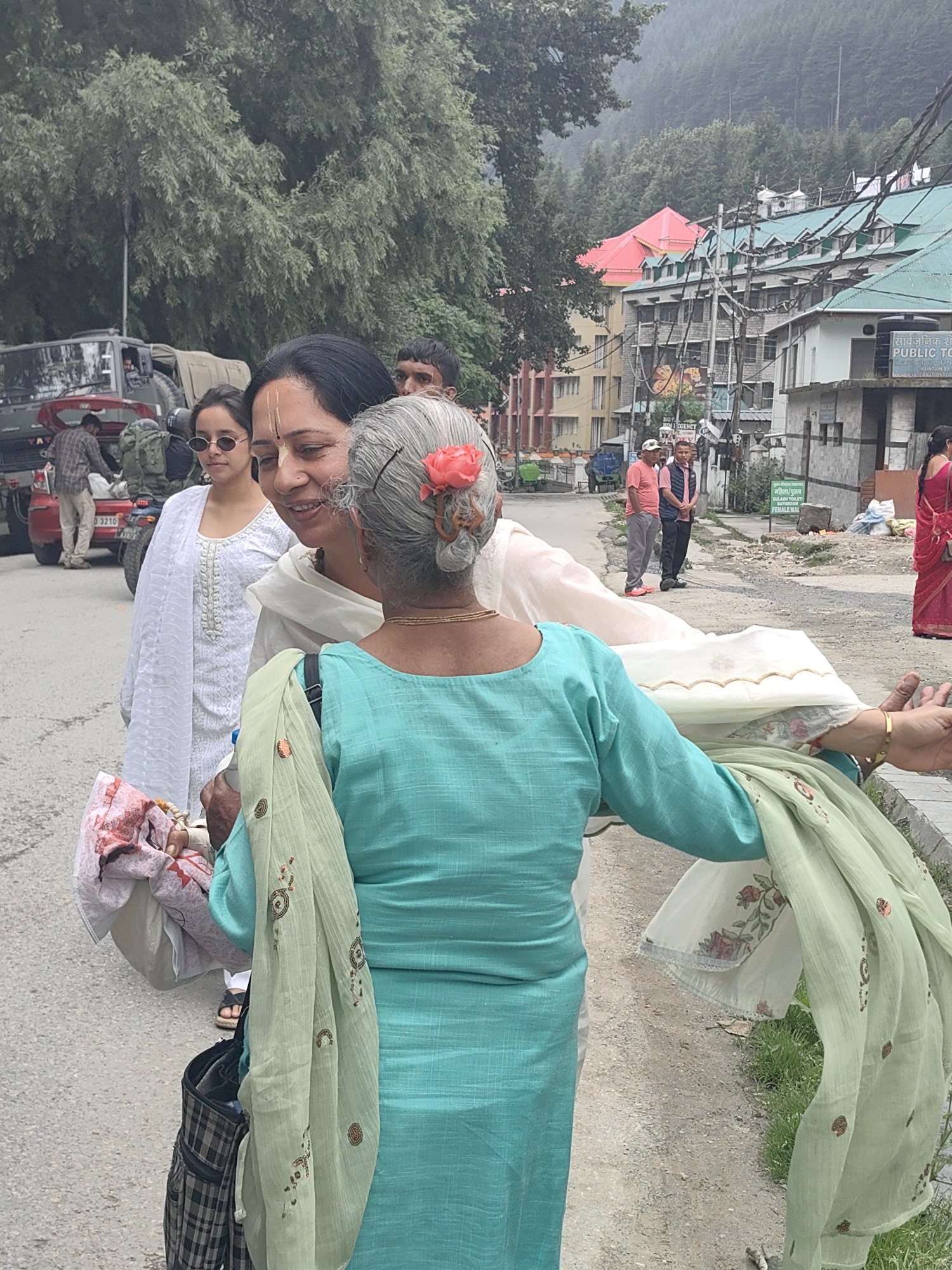 The image depicts a street scene in Manali, India, likely in a commercial area given the buildings in the background. The foreground is dominated by two women interacting. One woman, with gray hair adorned with a pink flower, wears a teal-colored kurta and holds a light green patterned scarf. She is facing away from the camera, towards another woman who is partially visible on the left. This second woman has dark hair, wears a white outfit, and has a traditional Indian religious marking on her forehead. She is looking at the first woman with a smile. Behind them, on the left, a young girl with dark hair and sunglasses stands, also looking towards the interaction. Further back, a parked military-style vehicle and a red car are visible, suggesting a busy or possibly tourist-oriented location. Buildings with green roofs and some with red roofs line the street in the background, indicating an urban or semi-urban environment. The street appears paved, and there is vegetation along the side. Several other people are visible in the middle ground and background, some walking and some standing. The overall atmosphere appears to be casual and social.