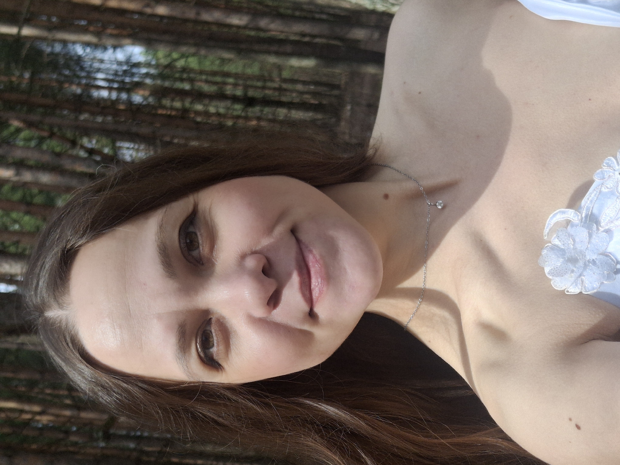 A woman with brown hair and fair skin is posing for a photograph in what appears to be a wooded area. The background suggests a forest with tall, thin trees, possibly pine or similar evergreens. The sunlight is filtering through the trees, creating patches of light and shadow on the woman's face and skin. She is wearing a light-colored garment with a decorative flower detail on the chest. A delicate silver necklace with a small pendant adorns her neck. Her expression is pleasant, with a slight smile. The location is not definitively determinable as Chaikovsky, Russia without additional contextual information.