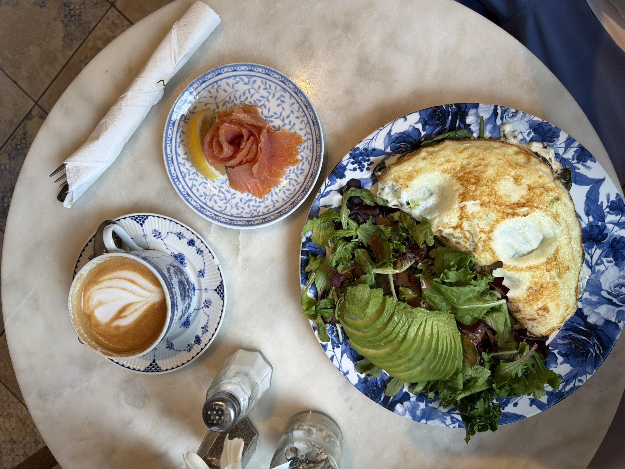 An overhead shot shows a marble tabletop with a breakfast spread in Miami Beach, United States of America. In the upper left, a cup and saucer with a latte art design sit next to a salt and pepper shaker. To the right, a white napkin is folded around silverware. In the upper right, a small plate holds smoked salmon shaped into a rose, with a lemon wedge. The main dish, on a large blue and white floral plate, features a salad with sliced avocado and an omelet topped with a dollop of cream. The scene is well-lit, highlighting the textures and colors of the food and tableware.