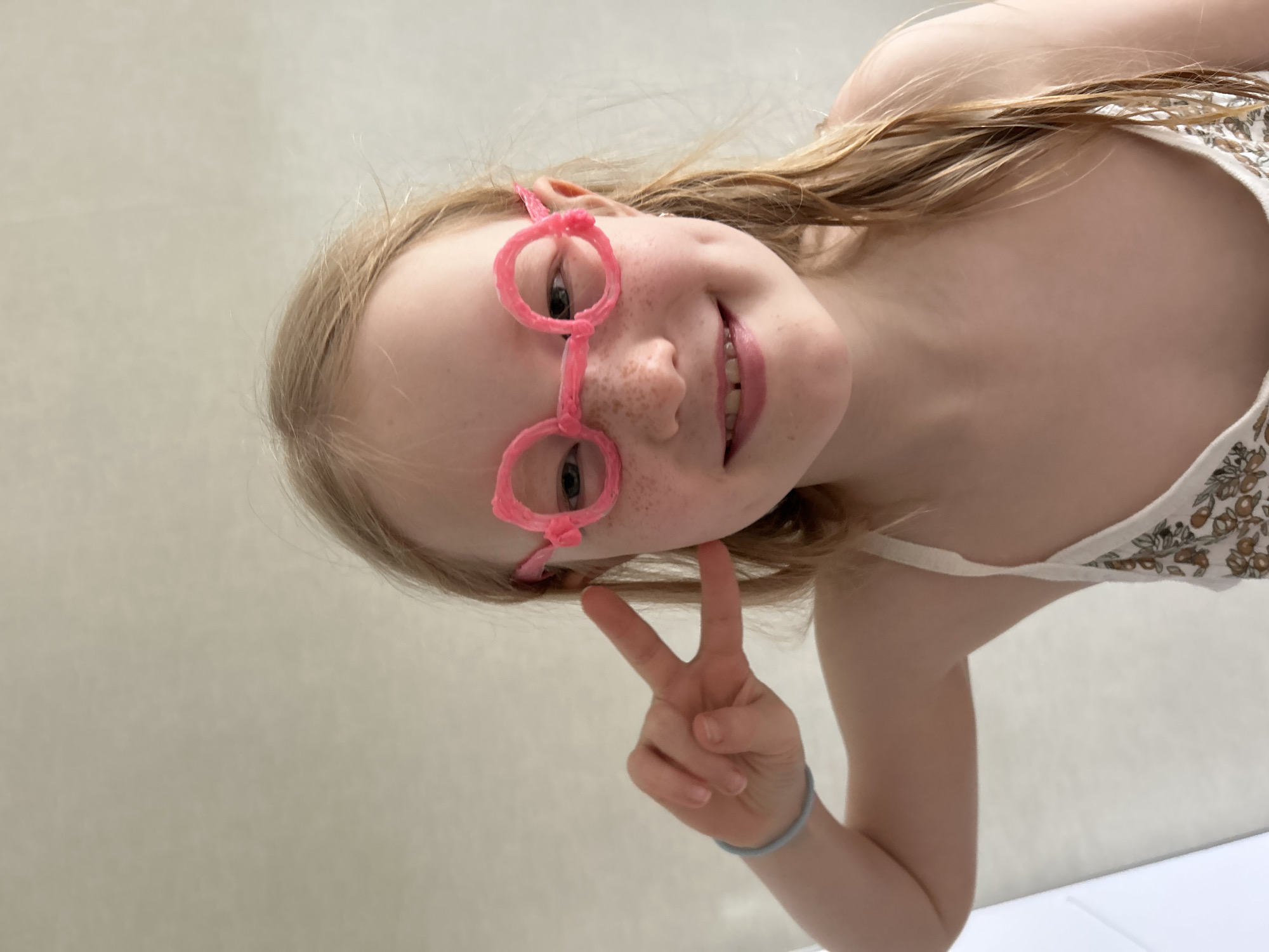 A young girl with blonde hair, freckles, and pink glasses is making a peace sign. She is wearing a light-colored floral top and a blue bracelet. The background is a plain, light-colored wall. The location is unknown and there is no indication of it being in Chaikovsky, Russia.