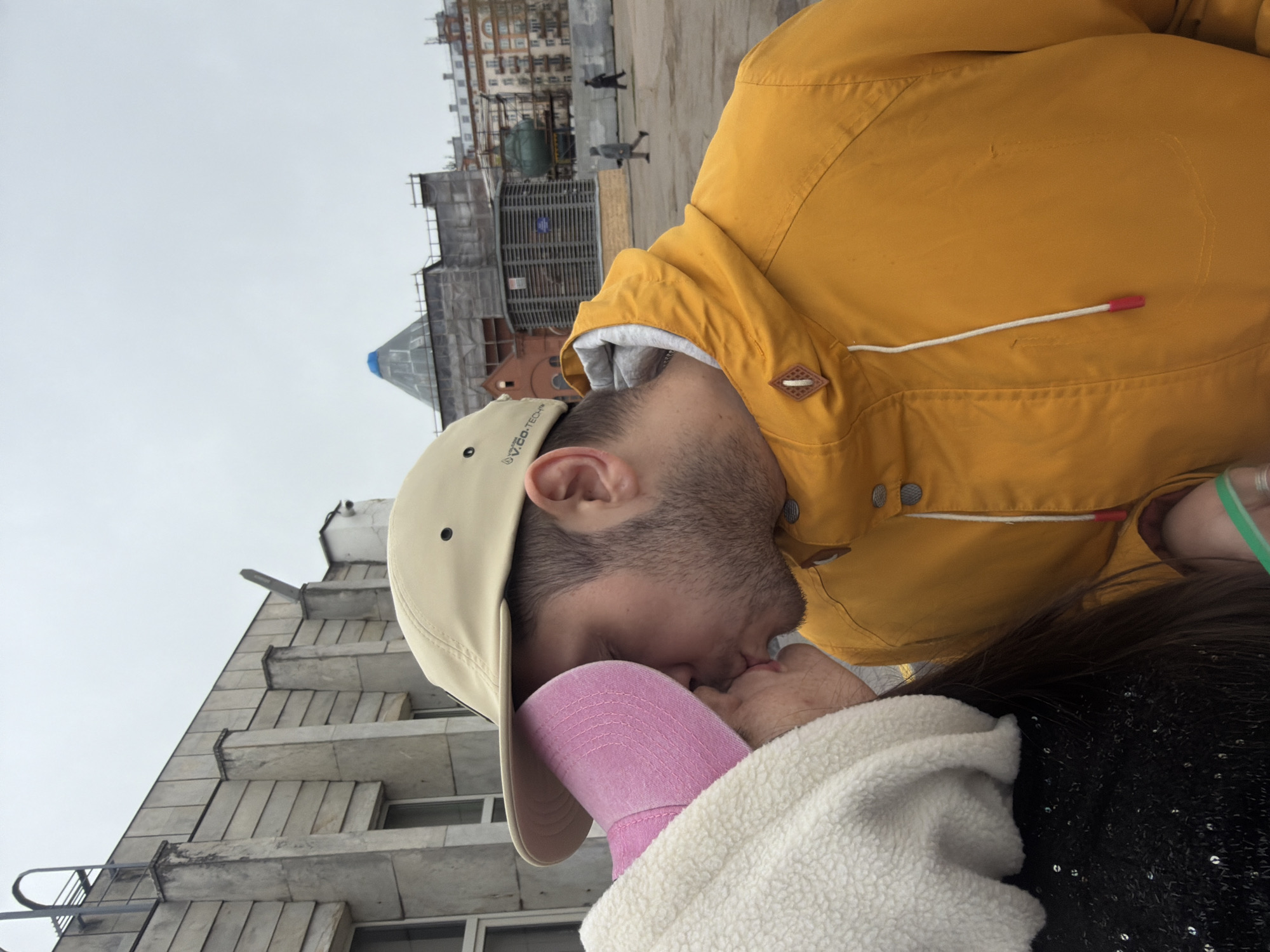 A couple is kissing in Saint Petersburg, Russia. The man wears a yellow jacket and a khaki baseball cap. The woman wears a pink hat and a white fuzzy jacket. The backdrop includes a building with scaffolding and a dome, and another building with stepped balconies. People are walking in the background. The sky is overcast.