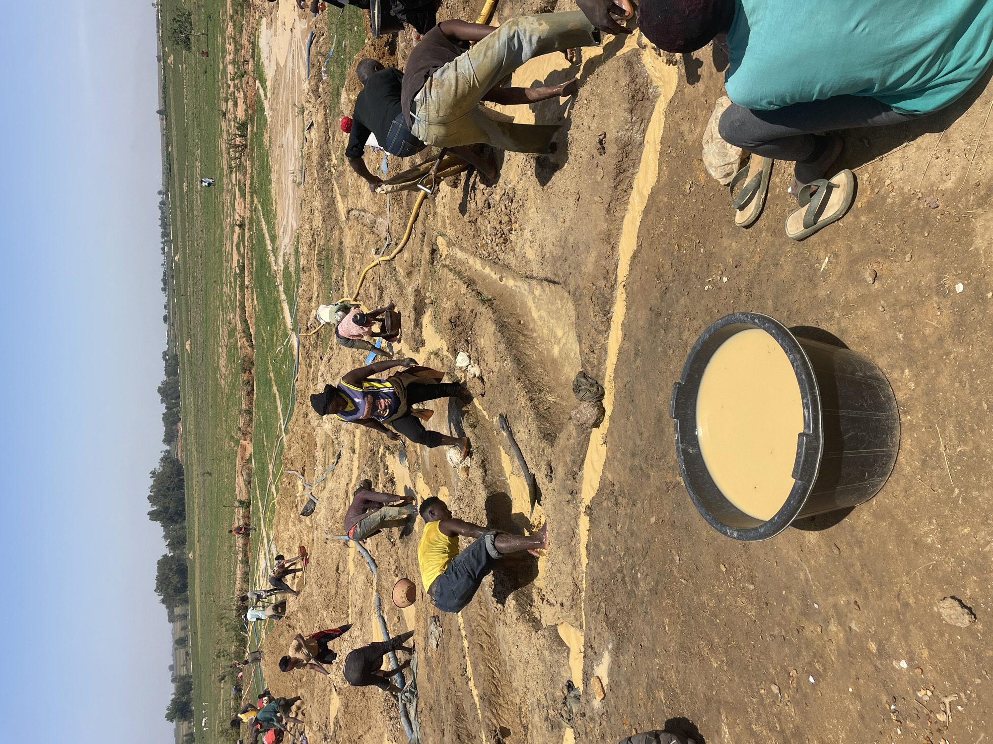 The image depicts a group of individuals working in a muddy area in Maiyanga, Nigeria. The scene is set on a field with a clear sky. A black bucket filled with a light brown liquid is in the foreground. Multiple individuals are seen digging and working in the mud, with some using tools. A yellow hose is visible on the ground. The environment appears to be a rural or agricultural area.