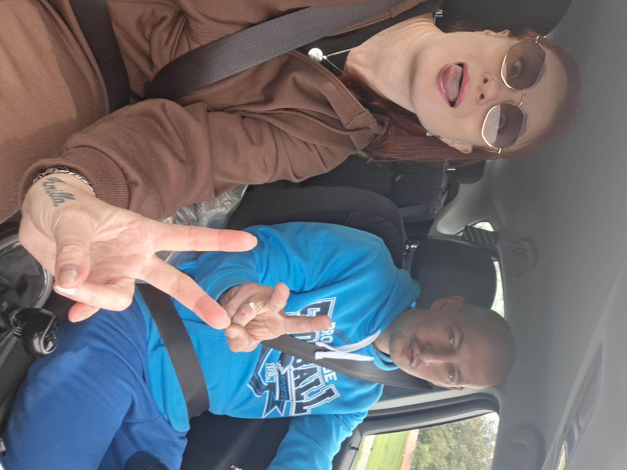 Two people, a woman and a man, are in the front seats of a car, appearing to be driving on a road in or near Myadzyel, Belarus. The woman on the left is wearing sunglasses and a brown hooded jacket. She has red hair and is sticking her tongue out, making a peace sign with her left hand. The man on the right, who is bald, is wearing a blue hoodie and blue pants. He is making a peace sign with his right hand and his left hand appears to be on the gear shift. The interior of the car is visible, including the dashboard, seats, and seatbelts. The exterior of the car shows trees and a grassy area through the windows, suggesting an outdoor, possibly suburban or rural setting, consistent with the outskirts of Myadzyel.