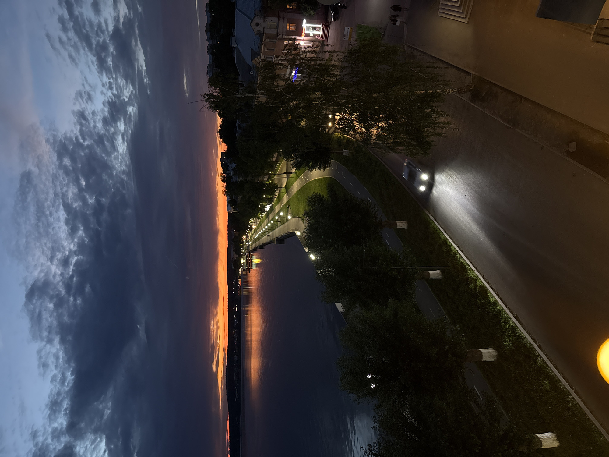 This is a vertical, high-angle, full shot taken at dusk in the city of Votkinsk, Russia, overlooking a lake and a street lined with trees and buildings. The sky is a dramatic gradient of dark blue and grey clouds tinged with fiery orange and yellow hues from the setting sun on the horizon. The lake, a deep blue, reflects the vibrant colors of the sky and the lights from the distant shore, where a line of buildings and streetlights can be seen. A paved promenade runs along the edge of the lake, illuminated by a row of lampposts. Adjacent to the promenade is a road with a bicycle lane. A car with its headlights on drives on the road, casting bright beams onto the wet asphalt. Trees, some with white-painted trunks, line the road and the promenade. On the right side of the image, there are multi-story buildings with windows and visible architectural details, suggesting an urban setting. Two people are walking on a sidewalk in front of these buildings. The overall atmosphere is serene and transitional, capturing the beauty of twilight in a Votkinsk cityscape.