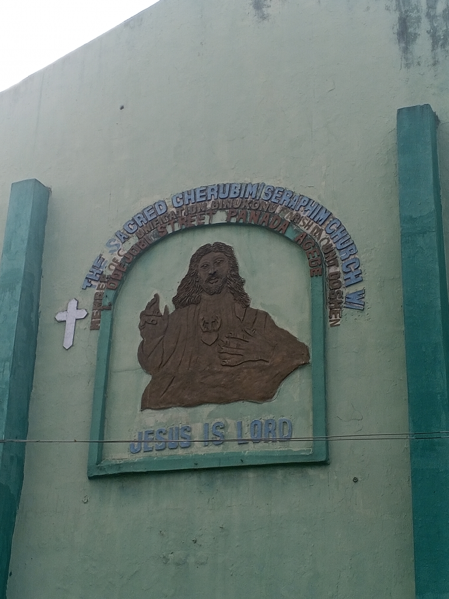 The image shows a religious building or church exterior in Jibowu, Nigeria. A green concrete wall features a mounted, textured depiction of Jesus Christ in brown. Above the image, arched text reads "THE SACRED CHERUBIM/SERAPHIM CHURCH W/ MEMBER OF C UNIFICATION BINUKONU PARISH MOUNT GOSHEN STREET PANADA AGEGE." Below Jesus, the words "JESUS IS LORD" are inscribed. A white cross is located on the left side of the image. The building appears to be weathered, with some discoloration and imperfections on the wall. Two greenish pillars stand on either side of the image and the building wall.
