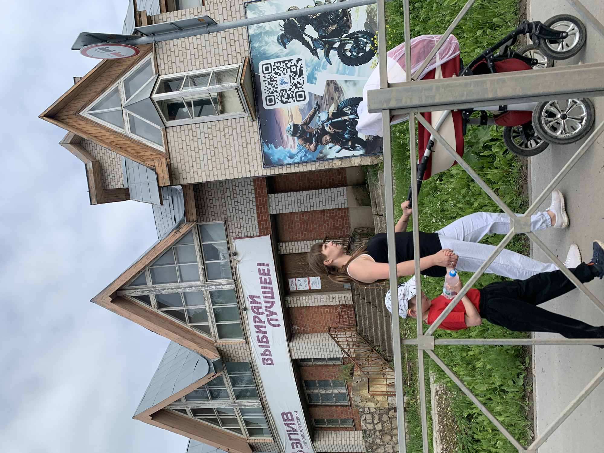 In Orda, Russia, a woman and a young boy walk past a building with a large banner that reads "ВЫБИРАЙ ЛУЧШЕЕ!" (Choose the best!). The building has a brick facade and distinctive triangular gables with wooden trim. To the right of the banner, a large advertisement featuring two men on motorcycles and a QR code is displayed. The woman is wearing a black tank top and grey pants, holding the hand of the boy, who is dressed in a red t-shirt, black pants, and a white cap. He is holding a bottle of water. Behind them, a red and white stroller is being pushed. A metal fence separates the sidewalk from a grassy area with weeds and some yellow wildflowers. The sky is overcast.