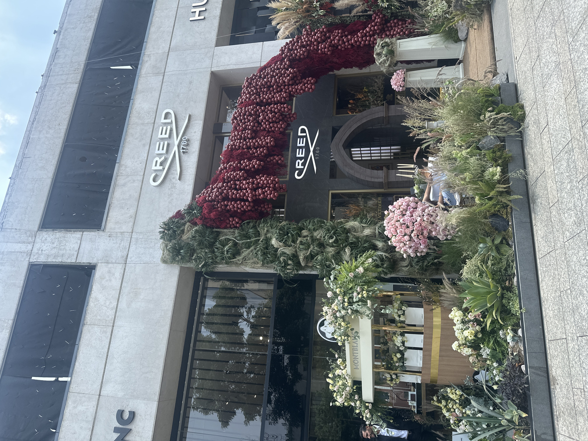 The image shows a commercial building facade, possibly a retail store, extensively decorated with floral arrangements. The building appears to be located in Mexico City, Mexico, as a Montblanc sign is visible. The main entrance is adorned with a large archway-like structure made of red and green floral elements. The storefront features the "Creed 1760" logo in white lettering. There is vegetation surrounding the entryway at ground level, including various types of flowers and grasses. The building has multiple windows, some of which are covered or obscured. There is a person partially visible amongst the flowers in front of the Montblanc sign. The image is taken from a slightly low angle on the sidewalk.