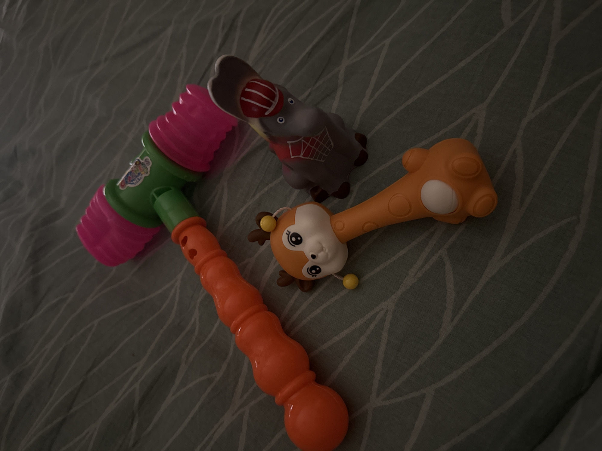 The image shows three toys lying on a gray, leaf-patterned surface. The toys include a pink and green hammer, a gray elephant with a basketball design, and a yellow and orange giraffe. The location appears to be indoors, possibly on a bed or similar surface.