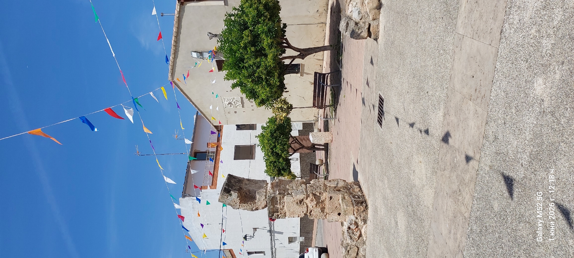 The image shows a street scene in Canals, Spain. The sky is clear blue. A string of colorful triangular pennants are strung across the street. The buildings are primarily white with some beige. There are two green trees on the right side of the image, next to a beige building with two benches. A stone pillar stands in the foreground. The ground is paved with gray concrete. The shadows of the flags can be seen on the ground. A white car is partially visible at the bottom. A camera watermark indicates the date: June 1, 2026.