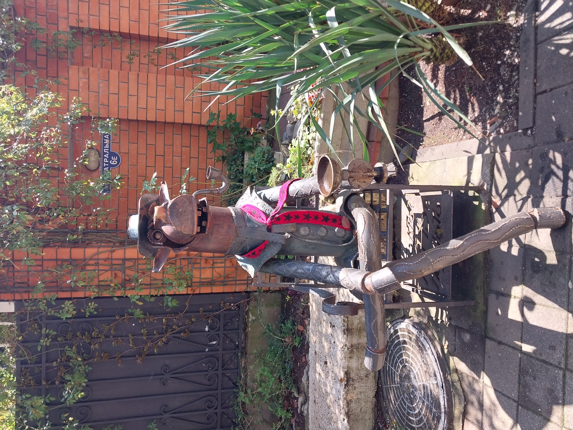 An outdoor, eye-level shot captures a whimsical metal sculpture set against a brick wall and lush greenery, likely in Sochi, Russia. The sculpture, crafted from repurposed metal parts, depicts an anthropomorphic figure resembling a creature or character, adorned with a hat and a red sash featuring black stars. It appears to be seated or mounted on a metal base, possibly an old industrial stand.
In the background, a warm-toned brick wall is partially covered by climbing vines and various green plants. A prominent plant with long, spiky leaves (likely a Yucca) stands on the right, casting distinct shadows on the paved ground. A black ornate metal gate or fence is visible on the left.
A blue street sign is attached to the brick wall, displaying the Cyrillic text "ТЕАТРАЛЬНАЯ" and the number "6Е". This identifies the location as "Teatral'naya Street" (Theater Street), building number "6E".
The scene is bathed in bright daylight, indicated by strong, crisp shadows, suggesting a sunny day. No direct activity is taking place; the image is a static portrayal of an outdoor art piece within an urban, green setting.