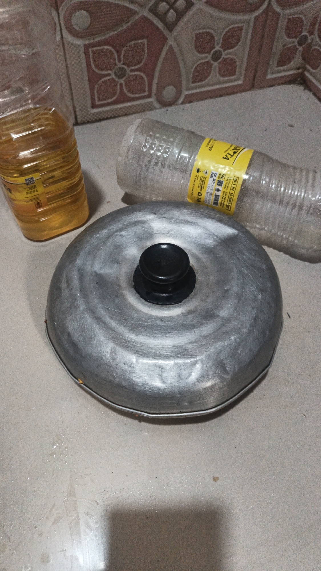 The image shows a top-down view of a metal bundt cake pan with a black knob in the center, placed on a light-colored, textured surface. To the left of the pan is a clear plastic bottle containing a yellow liquid, likely cooking oil, with a yellow label. To the right of the pan is another clear plastic bottle, partially obscured, which appears to be empty and has a similar yellow label with text and some white residue inside. In the background, there is a tiled wall with a repeating floral pattern in shades of pink and beige. The overall scene suggests a domestic setting, possibly a kitchen, and the lighting indicates it was taken indoors. There is no visible evidence to suggest this image is located in Kandangmas, Indonesia.