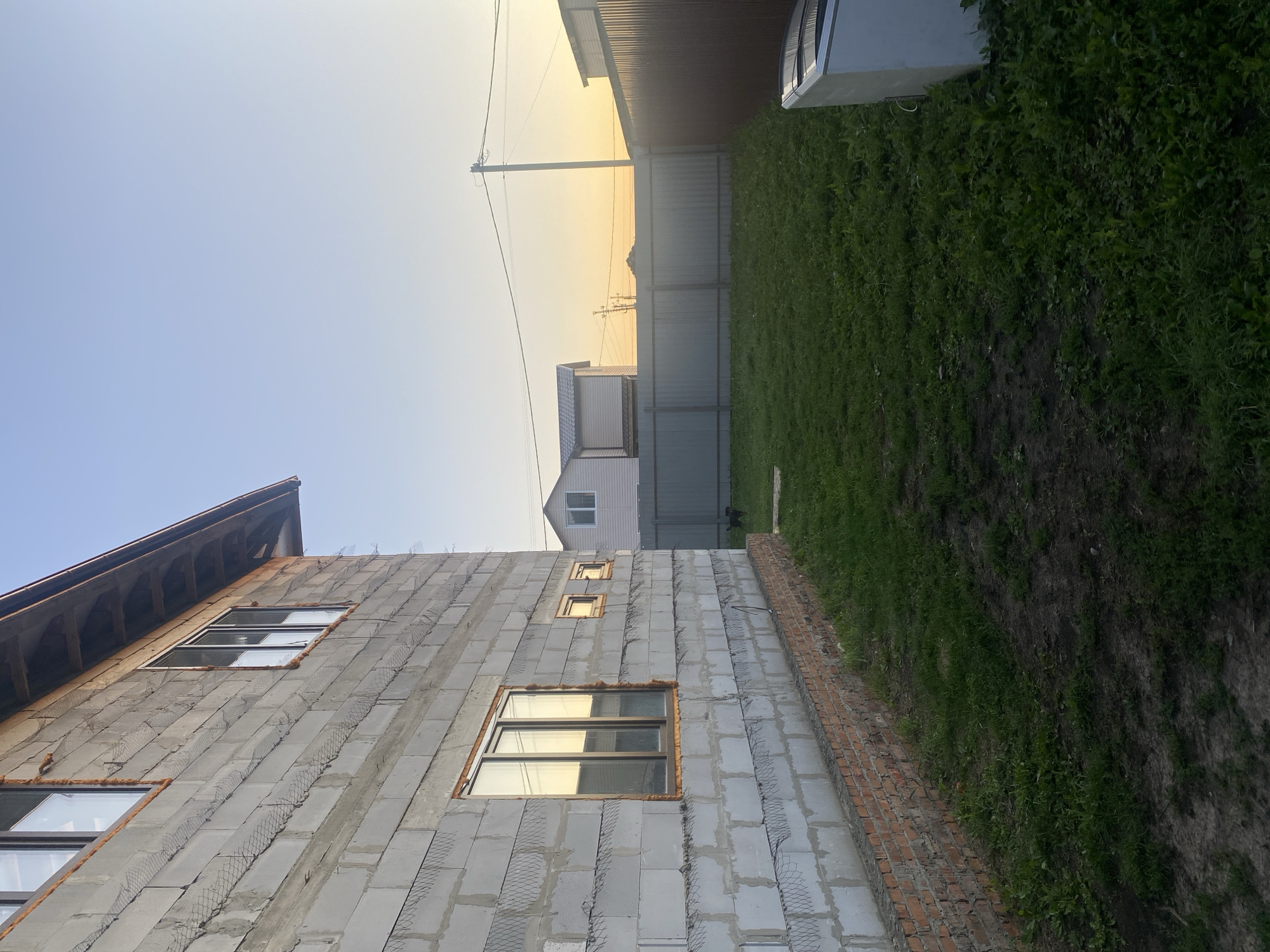 This image depicts the exterior of a two-story house under construction in the city of Verkhniy Karbush, Russia. The house is constructed from concrete blocks and features several windows, some of which are installed. A brick foundation is visible at the base of the building. The foreground consists of a grassy yard with some bare patches of soil. In the background, a metal fence separates the property from neighboring areas, beyond which are other houses and a utility pole. The sky is clear with a gradient of blue at the top and a soft yellow hue near the horizon, suggesting it is either dawn or dusk. A white appliance, possibly a freezer, is located on the right side of the yard. A small black dog is partially visible near the fence line.