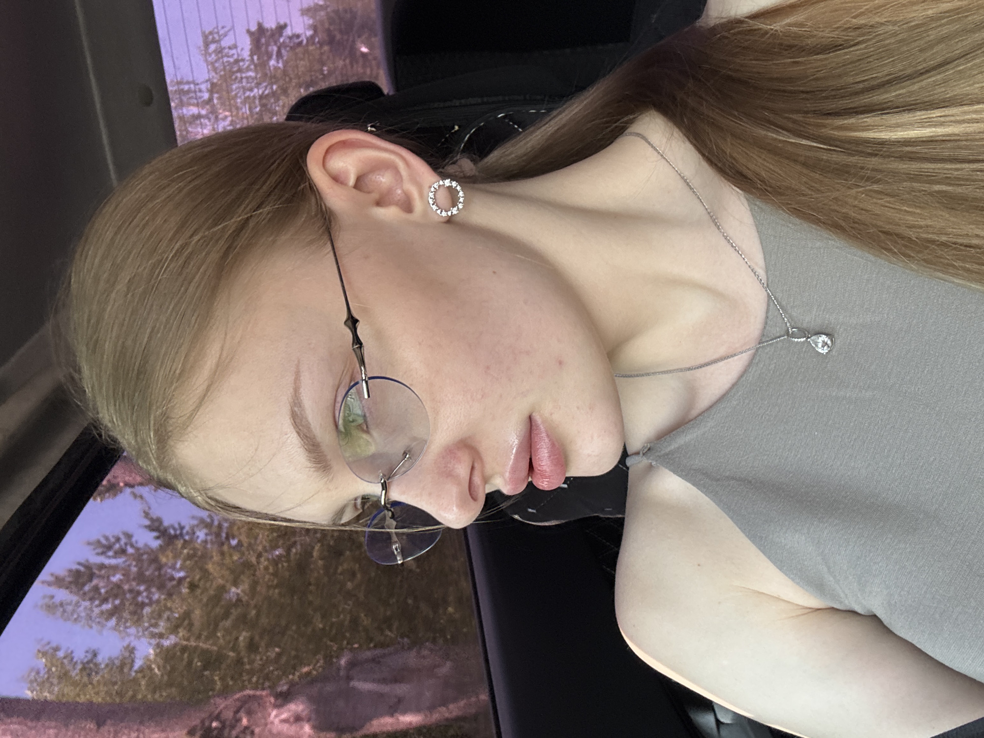 A fair-skinned young woman with long blonde hair is inside a vehicle, likely a car, as evidenced by the window frame in the lower left corner and the car seat behind her shoulder. She is wearing round, wire-framed glasses, a silver necklace with a teardrop pendant, and round, possibly diamond-studded, earrings. She is wearing a gray sleeveless shirt. Through the car window, trees are visible, and the light suggests either dusk or dawn. The location in Perm, Russia, is not explicitly discernible from the visual elements present.