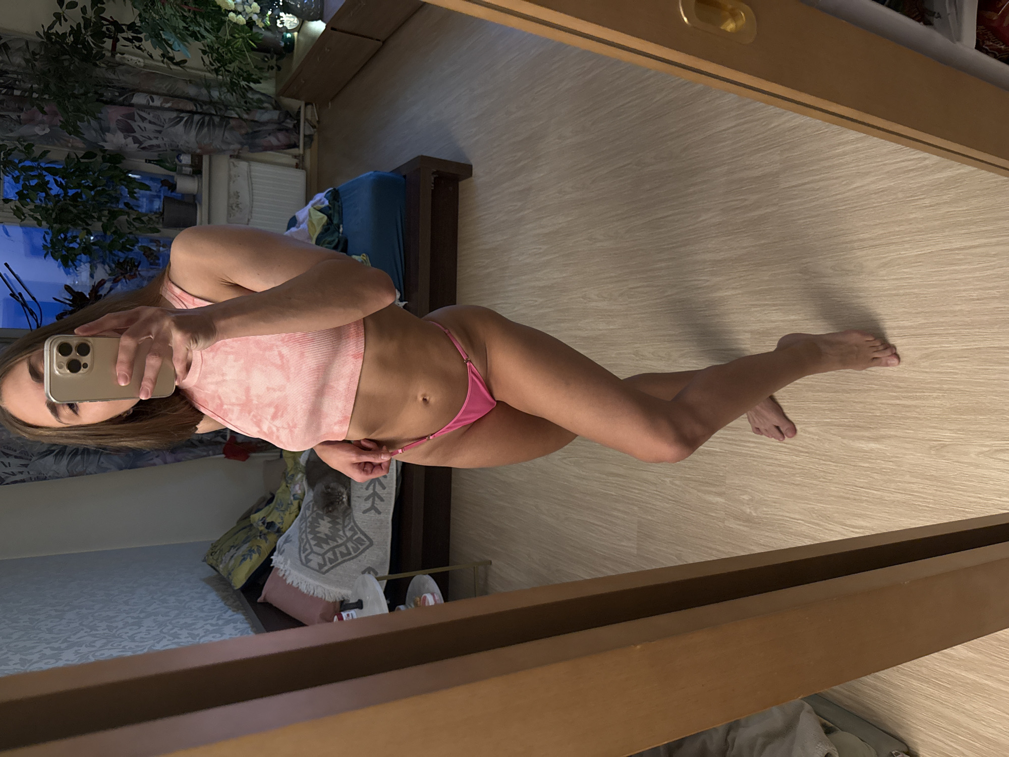 A mirror selfie captures a woman indoors, standing and posing. She is wearing a light pink, tie-dye or marbled ribbed crop top and hot pink bikini bottoms. Her face is obscured by a silver or light-colored smartphone with a multi-lens camera system, which she holds up to take the photo. Her long, dark hair falls over her shoulders.
The setting appears to be a bedroom or dressing area. The reflections in the mirror show a dark wooden bed frame with a blue upholstered headboard or mattress. A patterned rug with a geometric design is visible on the floor or draped over furniture. In the background, through a window, city lights or buildings are visible against a dark sky, suggesting it is evening or night. Floral patterned curtains hang beside the window, and a radiator or heating unit is also present. A green leafy plant adds to the decor. The floor is light-colored wood laminate. The mirror itself has a thick, dark wooden frame, and a gold handle is visible on the frame in the upper right. There is no visible text in the image.