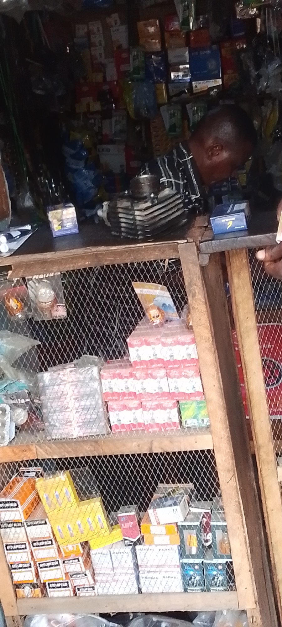 The image depicts the interior of a small shop in Lokoja, Nigeria. An older man is behind a wooden counter with a wire mesh front, looking at an engine part. Shelves behind the counter are stocked with various products, including boxed items and spare parts. The shop appears cramped and dimly lit.