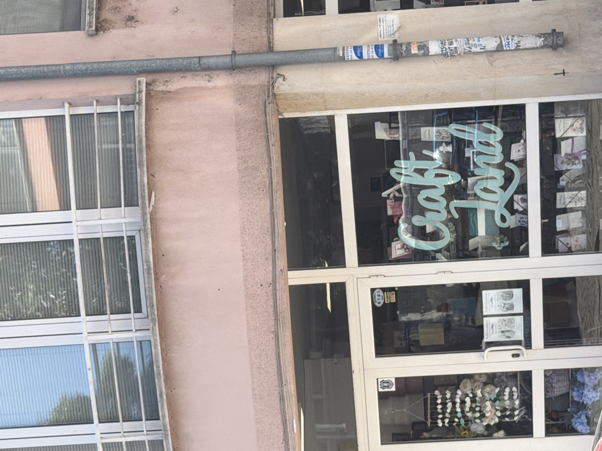 The image depicts the facade of a building in Plovdiv, Bulgaria, specifically a storefront identified as "Craft Land." The building has a window on the left, covered with metal bars. To the right, the storefront features a glass door and large glass windows displaying crafts and merchandise. A horizontal pipe runs along the upper part of the building facade. The overall impression is a small business situated in a possibly older urban environment, with potential signs of wear and tear on the building's exterior.