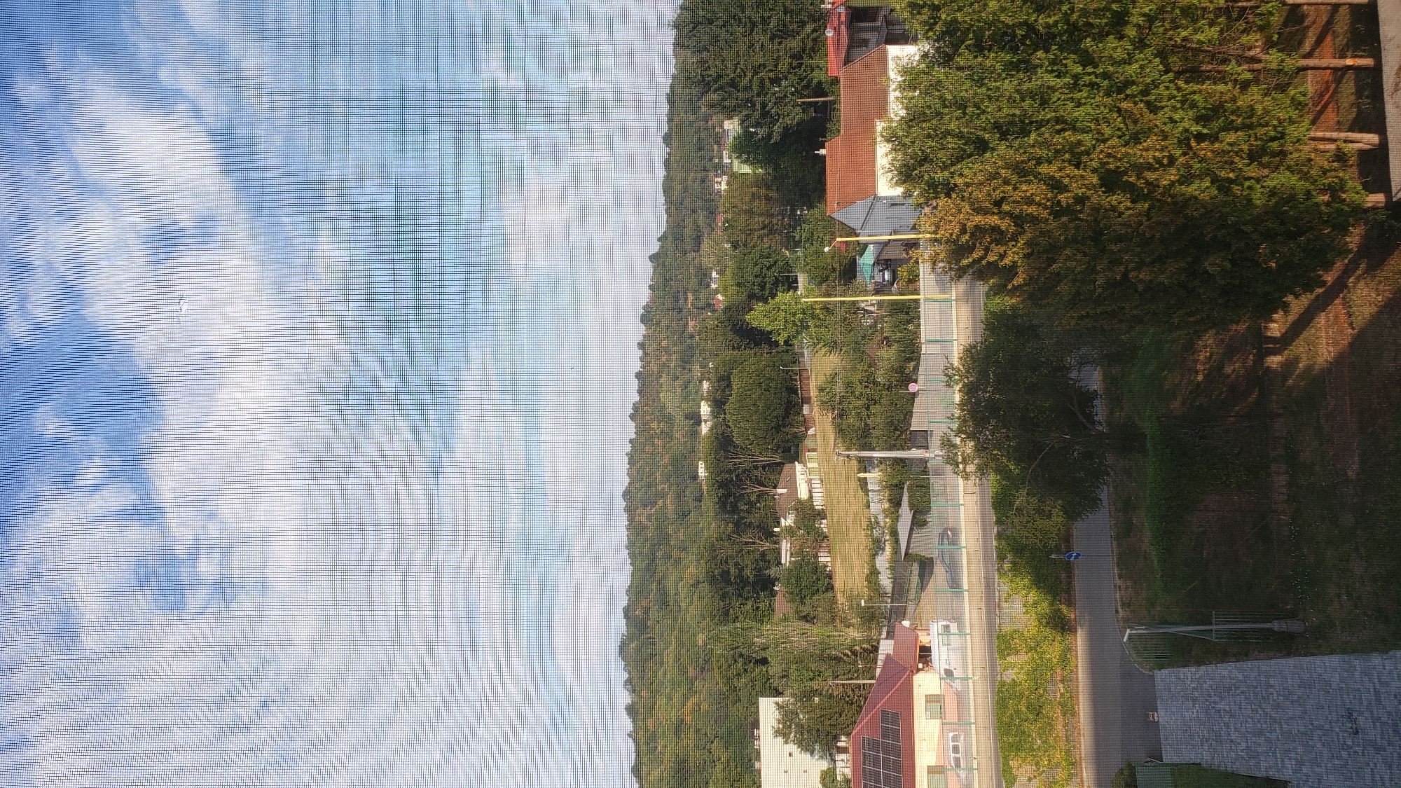 The image depicts a view from a window, showing a suburban landscape in Prešov, Slovakia. In the foreground, a grey paved walkway is visible, leading away from the viewer. Adjacent to it is a grassy area with sparse trees casting long shadows. A road runs horizontally across the mid-ground, with a fence separating it from properties on the other side. Several houses with red-tiled roofs are scattered amongst lush green trees, some with yellowing leaves, suggesting it might be late summer or early autumn. In the background, a densely wooded hill rises, covered in a variety of trees. The sky above is blue with scattered white clouds. The view is partially obscured by a fine mesh screen, creating a grid pattern over the entire image.