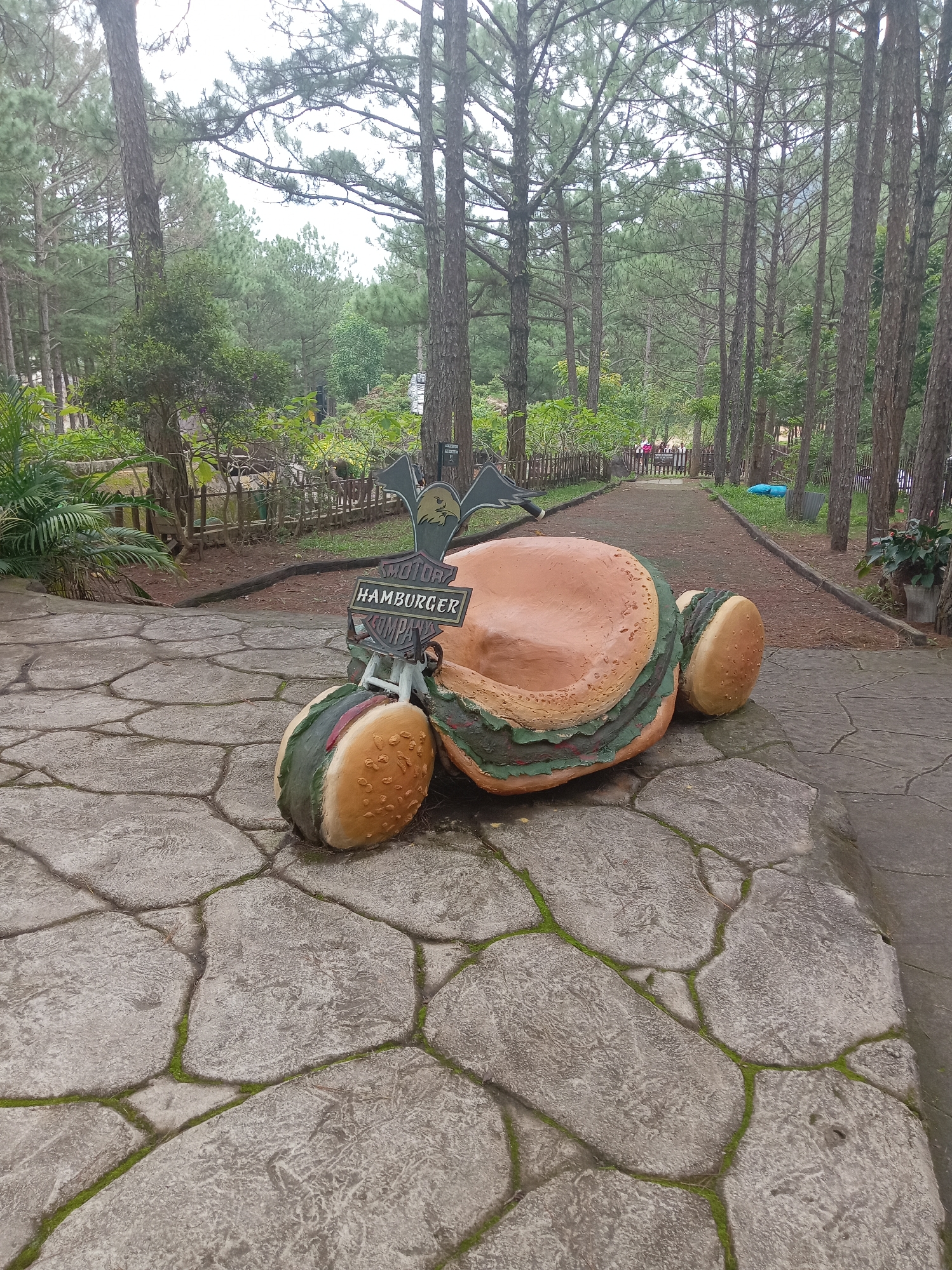 The image depicts a whimsical hamburger-shaped sculpture situated on a stone-paved path in a park-like setting. The sculpture, resembling a giant hamburger with buns, lettuce, and patty, is positioned on its side, with its bun-like wheels facing the viewer. A sign attached to the sculpture reads "MOTOR HAMBURGER COMPANY," suggesting it's an advertisement or a decorative piece for a food establishment.
The background reveals a pathway lined with tall pine trees, indicative of a natural or recreational area. The ground is covered with a mosaic of irregular-shaped stones, some of which have moss growing in the crevices. In the distance, more trees and foliage are visible, and a few people can be seen walking along the path, hinting at a public space. The overall ambiance is serene and outdoorsy.
This sculpture is located in Da Lat, Vietnam, specifically at the "Motorbike Burger" tourist attraction. It's known for its quirky, Instagrammable features, including this oversized hamburger sculpture. The location is characterized by its pine forests and cooler climate, making it a popular destination for both domestic and international tourists.