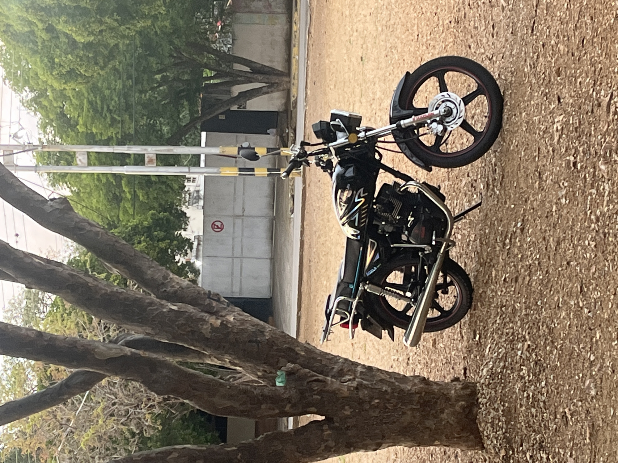 A black motorcycle is parked on a dirt ground next to a tree. The motorcycle has a chrome exhaust pipe and a red rim on its wheels. In the background, there is a concrete wall with a "P" sign. The scene appears to be in a public area with trees and buildings, possibly in Santa Ana, Venezuela.
