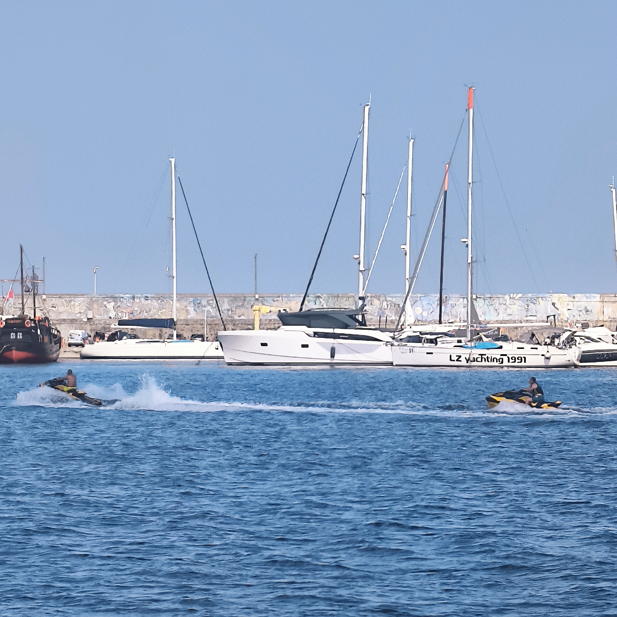 Two jet skiers are on the water in the foreground. The jet skier on the left is in motion, creating a spray of water. The jet skier on the right is also in motion, with less visible spray. In the background, several sailboats are docked. The largest sailboat on the right has "LZ Yachting 1991" written on its hull. To the left of the sailboats, a dark-colored boat with red trim is docked. Behind the boats, a concrete wall with graffiti is visible. The scene appears to be a harbor or marina, likely in Varna, Bulgaria, given the presence of sailboats and the general coastal environment. The water is blue and choppy, suggesting a breezy day. The sky is clear and blue.