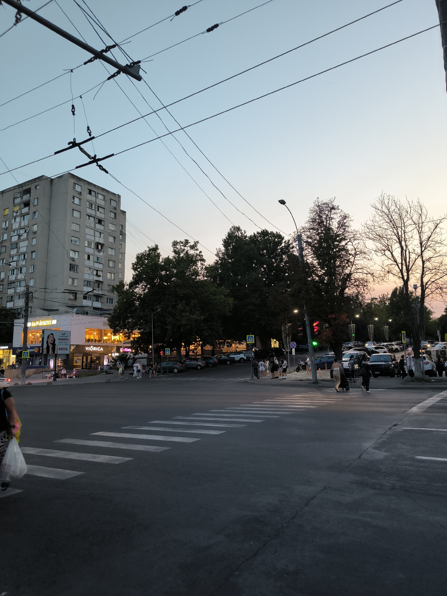 The image depicts a street scene at dusk in Chișinău, Moldova. The foreground shows a pedestrian crossing on an asphalt road. In the midground, there is a busy intersection with several cars and people. Traffic lights are visible, showing green and red signals. A multi-story apartment building with a weathered facade and numerous windows stands prominently on the left side of the frame, with some balconies visible. Across the street from the apartment building, there are commercial establishments with illuminated signs, including "VIORICA" and another sign that appears to be "Restaurant." Trees line the street, with dense foliage on the right and bare branches on the left, suggesting a change of seasons. Overhead, a complex network of trolleybus wires crisscrosses the sky. The sky exhibits a gradient of pale blue to a light orange and pink hue, indicating the end of daylight. Several individuals are walking on the sidewalks and crossing the street, some carrying bags. The overall impression is a lively urban environment during the transition from day to night.