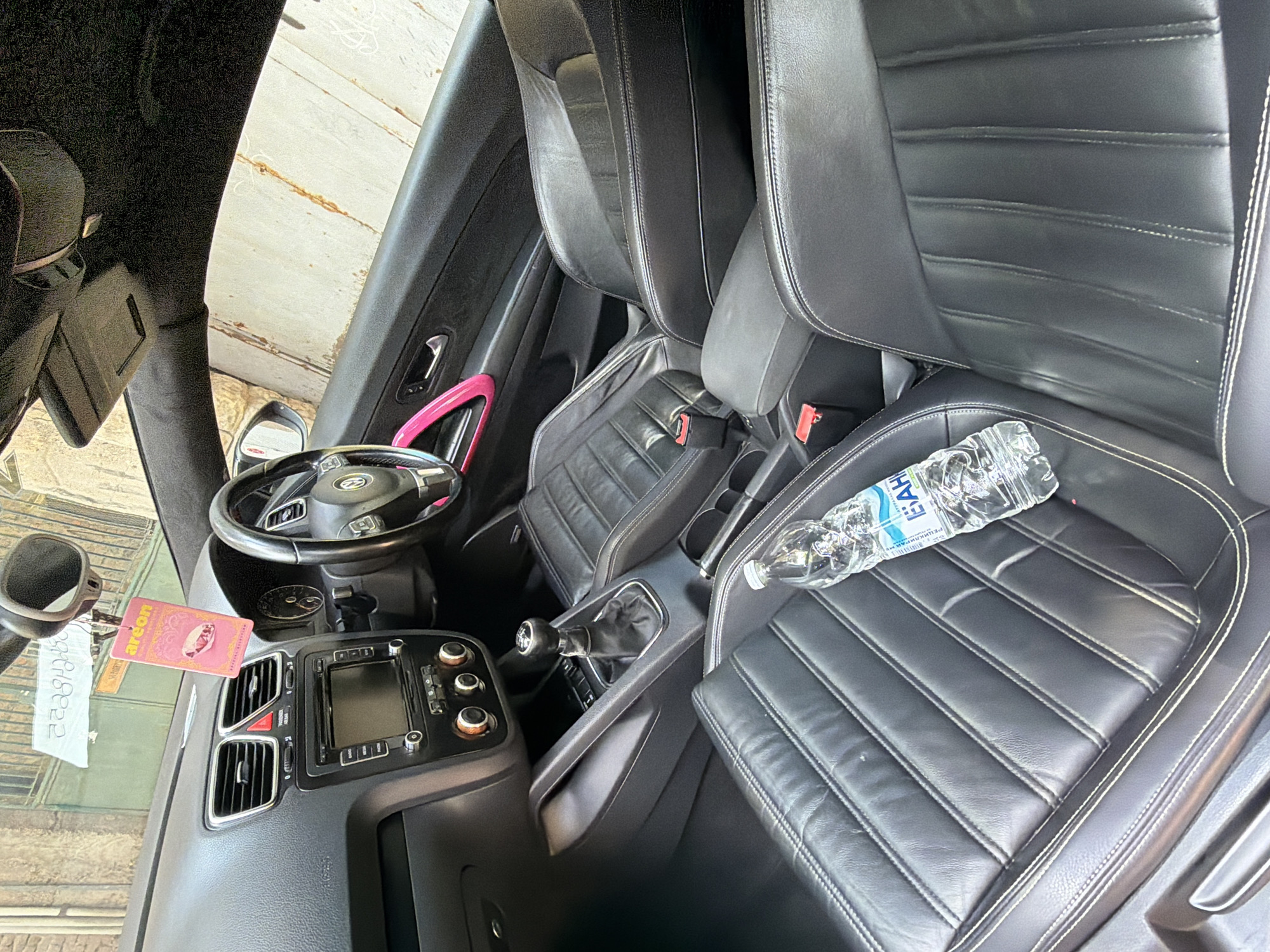 The image shows the interior of a car, specifically the driver's side and passenger seat. The seats are black leather with white stitching. A bottle of water with a label in Cyrillic script sits on the passenger seat. A pink air freshener is hanging from the rearview mirror. The car appears to be parked outdoors, with a glimpse of a building and what looks like a garage door in the background. The location is Plovdiv, Bulgaria. The scene is static and no people are visible.