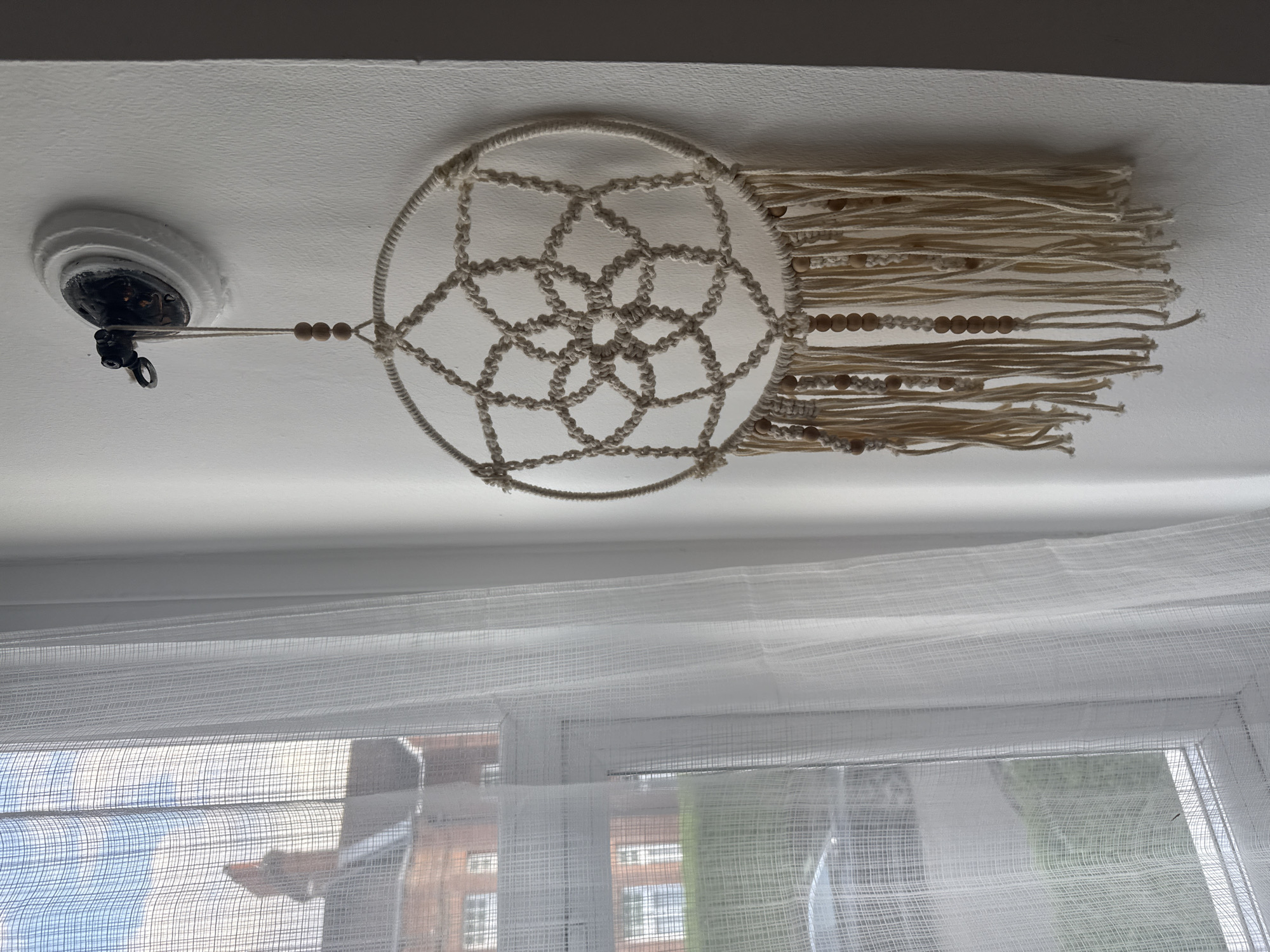 A dreamcatcher hangs from the ceiling in front of a window. The dreamcatcher consists of a circular frame, a woven web, and tassels with wooden beads. It is attached to a light fixture on the ceiling with a cord and wooden beads. The window has white sheer curtains. Outside the window, a brick building is visible. The scene is likely indoors during the day. Given the urban architecture, this could be a location in London, United Kingdom.