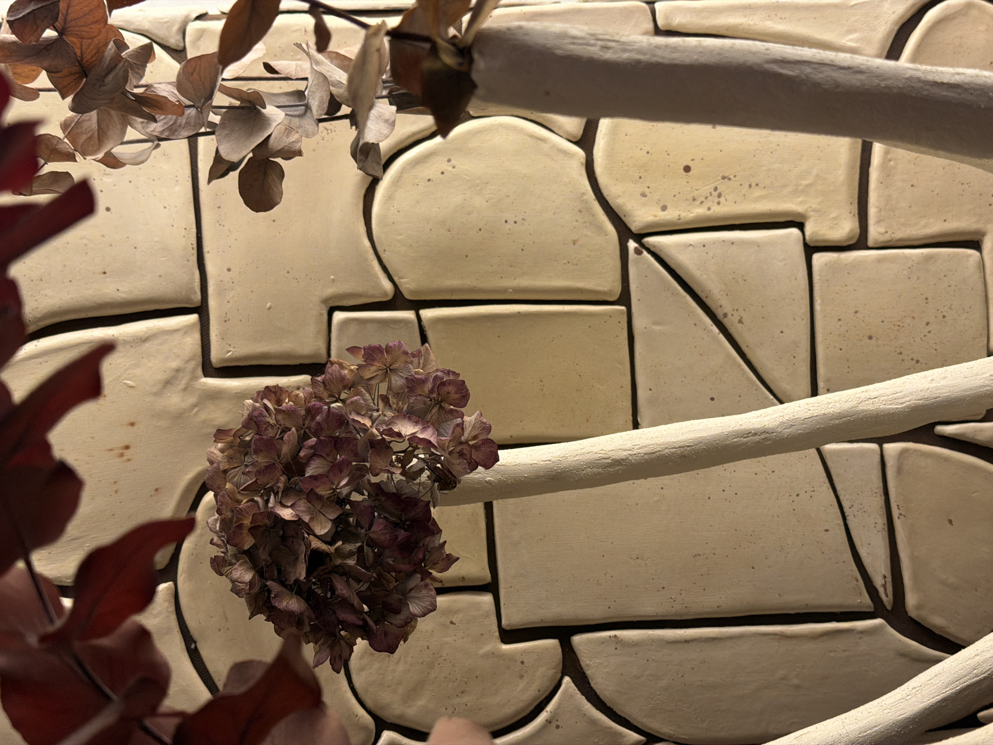A close-up shot of a decorative wall or display. The wall is composed of light beige or cream-colored tiles with irregular shapes and dark grout lines. A dried hydrangea flower head, predominantly purple and brown in color, is attached to the wall on the left side of the frame. Two off-white branches or rods are placed diagonally across the tile wall. Some dried leaves are visible near the top left corner, and other dried red leaves are in the bottom left corner of the frame. The lighting is soft and warm, giving the scene a rustic or vintage feel. Location information (Paris, France) is not visually discernible.