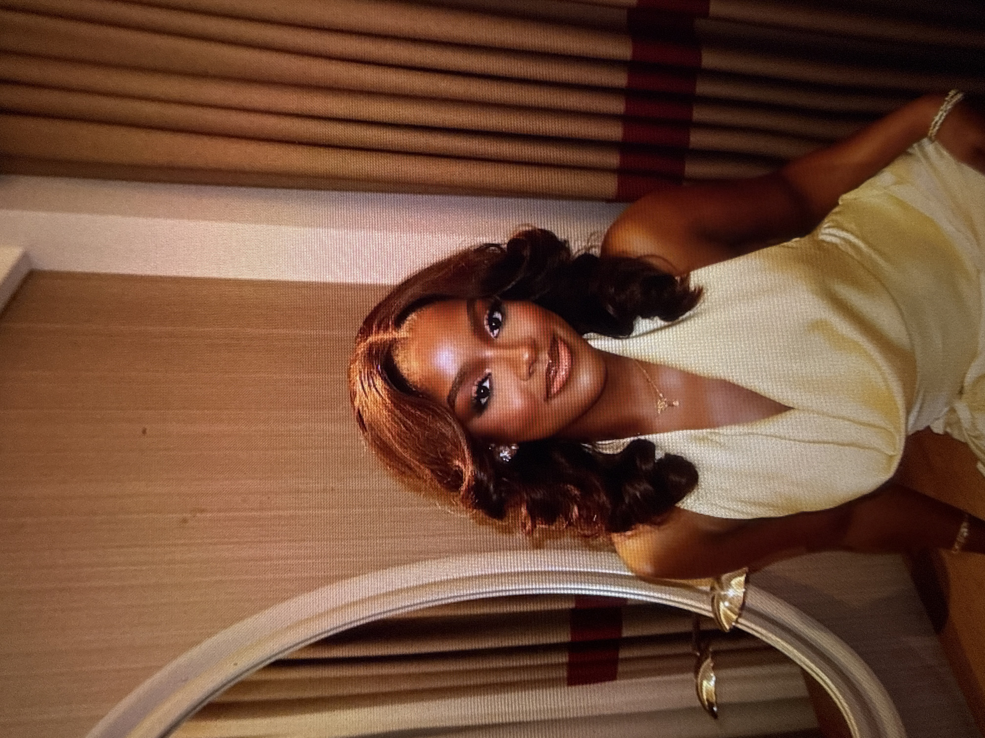 A young Black woman with dark, voluminous, wavy hair styled with a middle part is posing for a portrait in what appears to be a hotel room. She is wearing a light yellow, halter-neck dress with a deep V-neck and a draped bodice. She has on noticeable makeup, including eyeshadow, eyeliner, mascara, and lip gloss. She is adorned with earrings, a delicate necklace, and a bracelet on her right wrist. The background consists of textured tan curtains with horizontal stripes of maroon and cream, and to her left, a large, ornate oval mirror with a silver frame is visible, reflecting part of the room. The overall impression is one of elegance and glamour. The provided hints do not offer any information regarding the location within Abuja, Nigeria.
