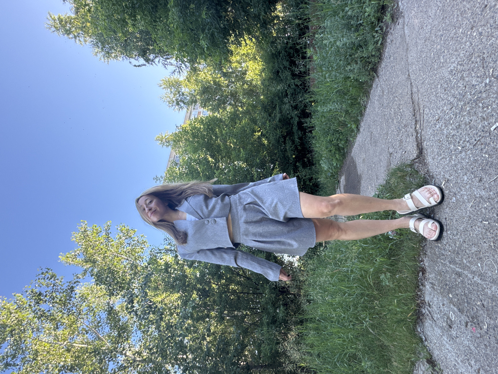A young woman stands on a paved path outdoors. She is wearing a grey cropped blazer over a white top and matching grey pleated shorts. She has on white sandals. The path is surrounded by lush green grass and trees. In the background, a light-colored building with windows is visible. The sky is clear and blue. The image appears to have been taken in Chaikovsky, Russia.