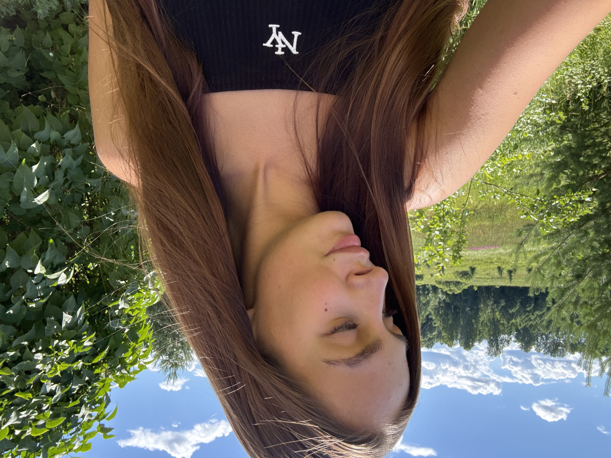 A young woman with long, brown hair is photographed outdoors in a rural setting, likely on a sunny day. She is wearing a black tank top with a white "NY" logo. Her head is tilted down and to her right, with her eyes closed. The background consists of lush green trees, fields, and a bright blue sky with scattered white clouds. The location is described as the city of Sholokhovo, Russia.