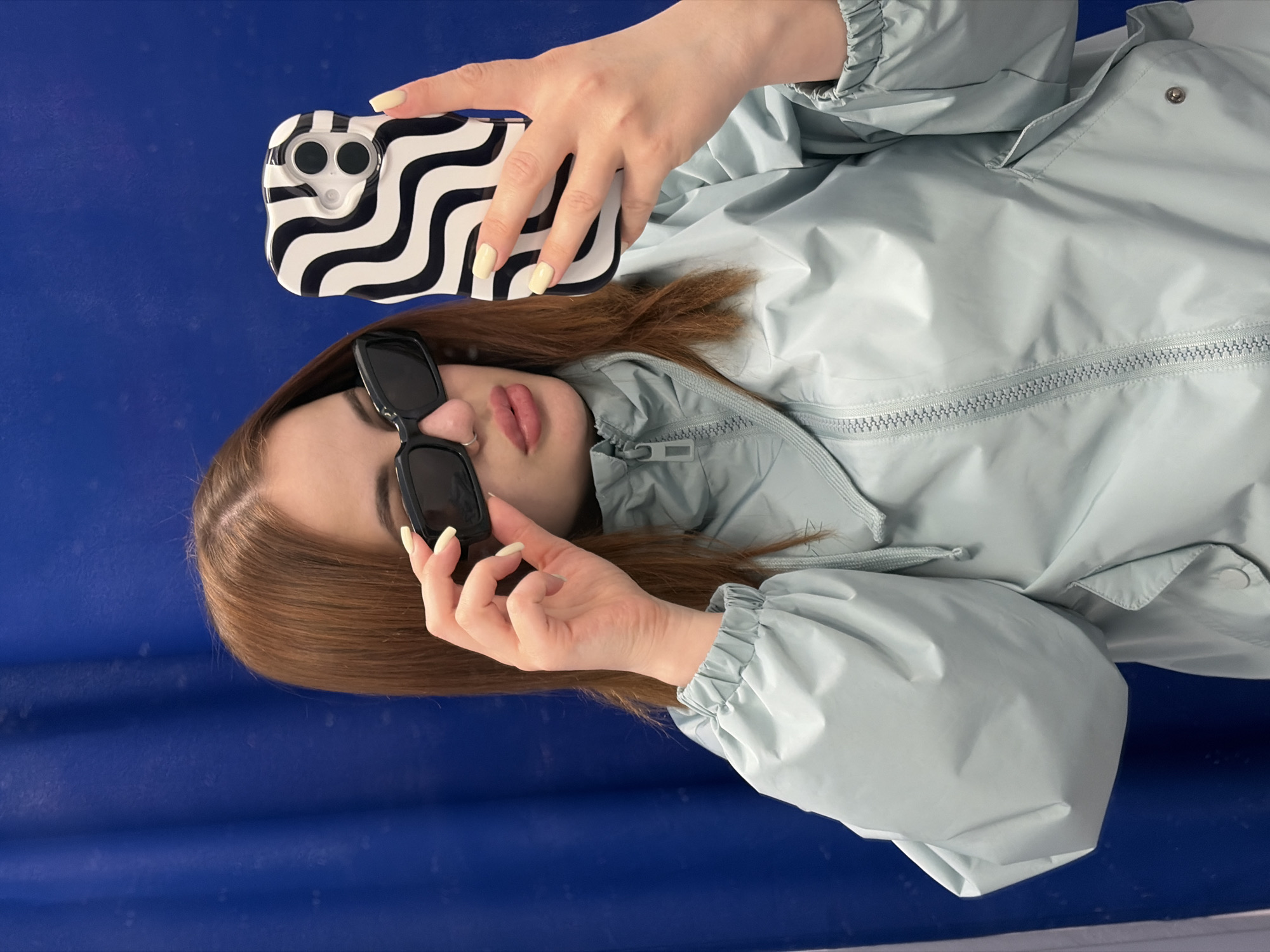 A young woman lies on a blue surface, taking a selfie with her smartphone. She is wearing a light blue jacket and black sunglasses. Her hair is brown. The phone case is black and white with a wavy pattern. The background is a plain blue surface. The location is unknown.