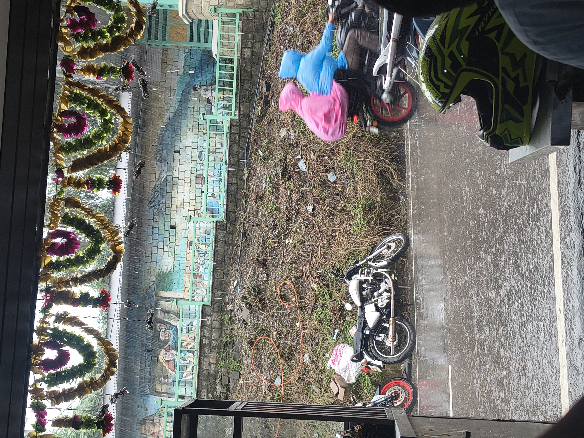 The image depicts a scene in Dharamshala, India, during heavy rainfall. A mural adorns a brick wall in the background, depicting figures and scenes. Festooned garlands hang overhead, adorned with floral elements. On the wet asphalt road, two motorcycles are parked. One is a white Royal Enfield, the other is a partial view of a motorcycle with a red rim. Further down the road, two individuals are riding a motorcycle, both wearing rain ponchos - one pink, the other blue. The ground is covered with dry grass, twigs, and some scattered debris. A green metal railing separates the road from the embankment. In the foreground, a brightly colored helmet with a green and black pattern rests on a metal surface. The overall atmosphere is that of a rainy day in an urban or semi-urban setting.