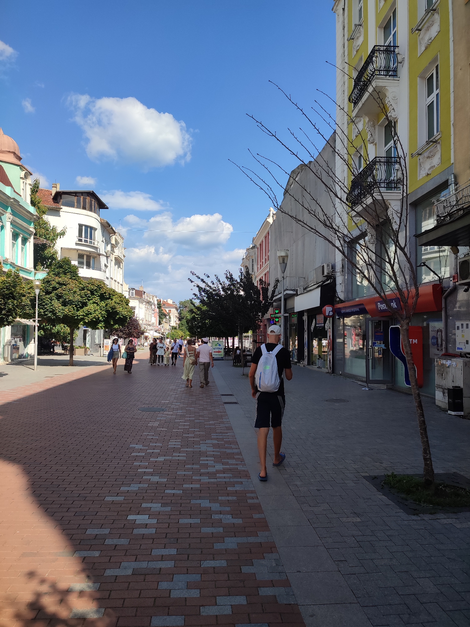 The image depicts a street scene in Varna, Bulgaria, likely on a sunny day. A pedestrian walkway paved with red and grey bricks dominates the foreground. The walkway is lined with buildings on both sides. On the left, there are multi-story buildings with classical architectural elements, including ornate balconies and arched windows, painted in light colors like white and turquoise. Lush green trees are planted along this side of the street, providing shade.
On the right, the buildings are also multi-story, with one prominent building painted in a bright yellow hue, featuring balconies with black wrought-iron railings. Shops and businesses are visible at street level, with signage including "Postbank" and what appears to be a pharmacy. A bare-branched tree stands on this side, casting a shadow on the walkway.
Several people are walking along the pedestrian area, some in small groups and others individually. The individuals are dressed in casual summer clothing. In the foreground, a young person wearing a white cap, a backpack, dark shorts, and flip-flops is walking away from the camera towards the right side of the street. The sky above is a clear blue with scattered white clouds. The overall impression is of a lively urban environment.