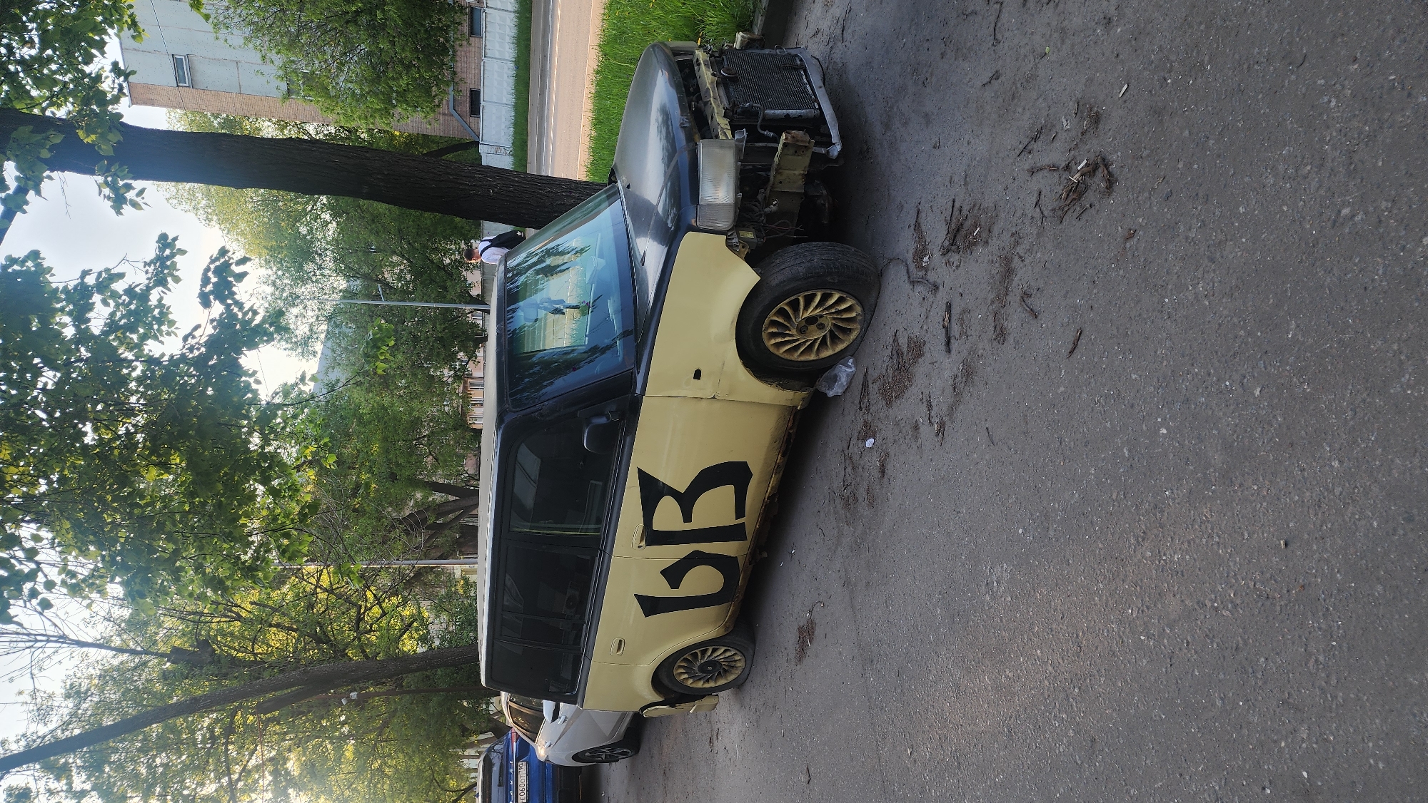 A heavily damaged yellow and black minivan, displaying the Cyrillic letters "БВ" on its side, is parked on a city street. The front end of the vehicle is significantly damaged, with the radiator exposed and the front bumper missing. The tires are yellow and appear to be deflated. The scene is outdoors, likely in a residential area with trees lining the street. Other parked vehicles are visible in the background, including one with a Russian license plate. The location is Moscow, Russia. There is a person in the upper portion of the photo with a white shirt.