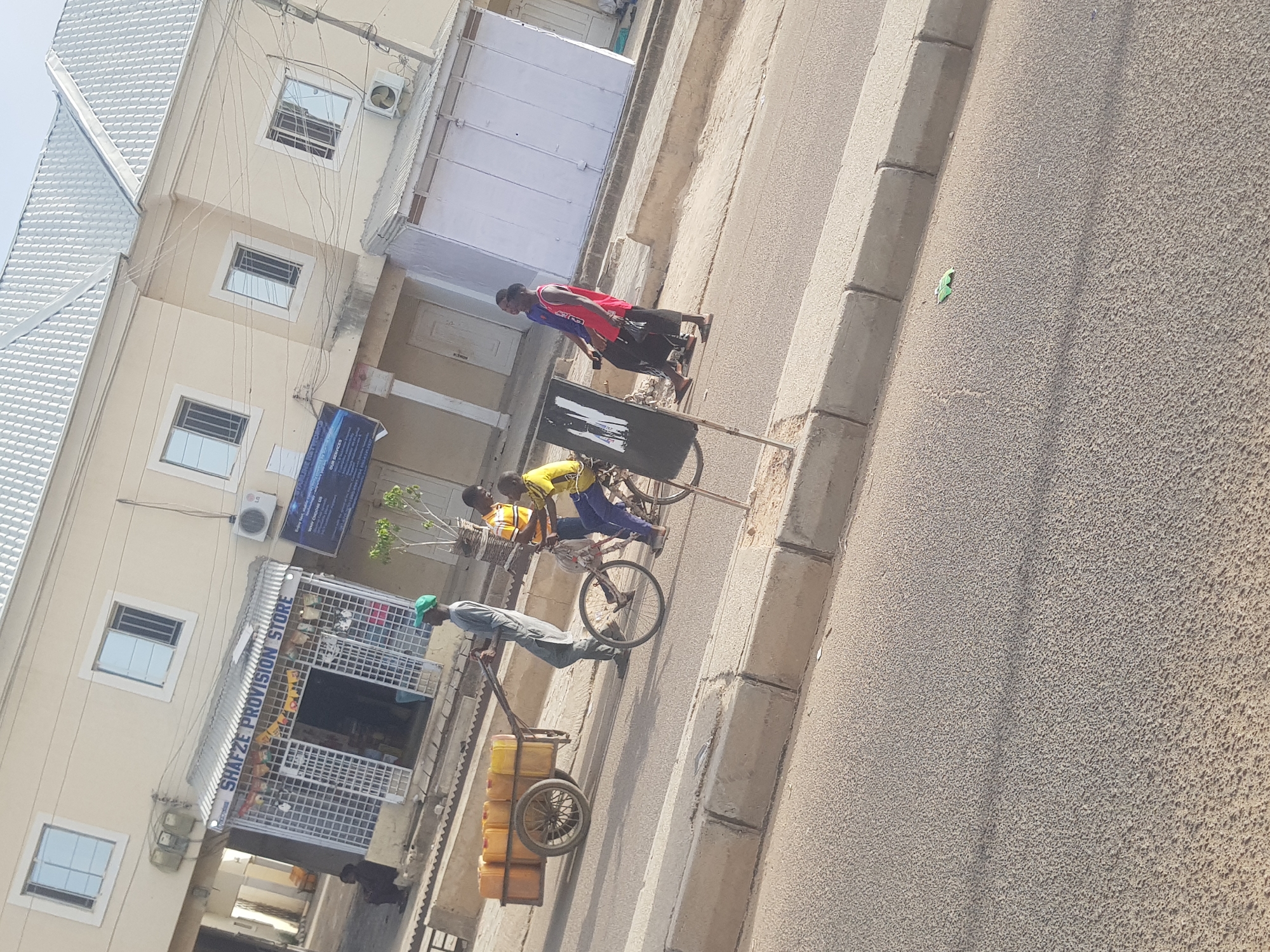 The image shows a street scene in what appears to be Dabawa, Nigeria. A man wearing a green cap and light clothing is pulling a two-wheeled cart loaded with yellow jerrycans. Two boys are riding a bicycle near him, while another boy is walking near the street. The setting includes a building with "SHAFZE PROVISION STORE" sign. The location appears to be a commercial street. The environment is bright and sunny, and the street is paved.