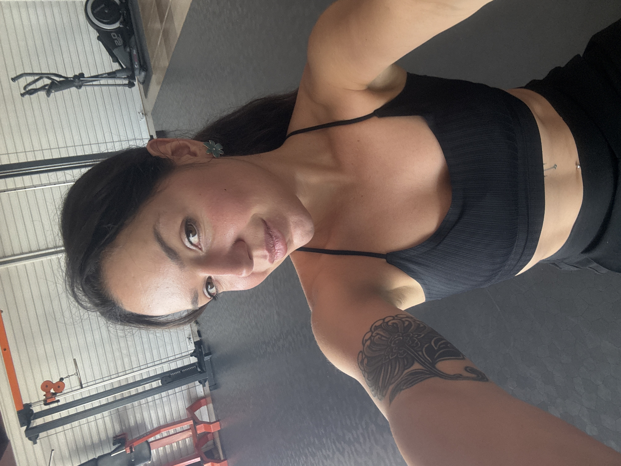 A woman with dark hair and a tattoo on her left arm is taking a selfie in a gym. She is wearing a black sports bra and black shorts. In the background, there are exercise machines, including a cable crossover machine and an elliptical trainer. The gym floor is covered with black mats. The gym appears to be located in Burgas, Bulgaria, based on the context provided.