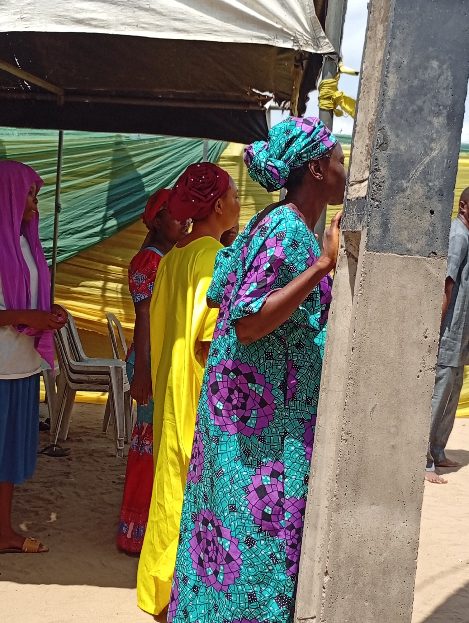 Several women stand near a concrete pillar under a tent in Adankolo, Nigeria. The woman closest to the pillar wears a teal and purple patterned dress and a matching headwrap. The next woman wears a yellow dress with a red head covering. Another woman is in red. To the left, a woman wears a purple hijab, a white shirt, and a blue skirt. A few white chairs stand in the background behind the women. A man wearing gray pants and no shoes stands partially visible to the right of the pillar. Green and yellow fabric hangs from the tent behind the women. The ground appears to be sandy.
