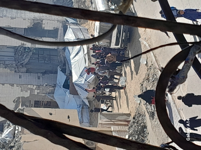 The image depicts an outdoor scene in Khan Yunis, Palestinian Territory, viewed through dark, out-of-focus metal foreground elements. The environment is characterized by extensive structural damage to multi-story buildings in the background, exhibiting collapsed sections, exposed interiors, and visible debris. Numerous white tarp-covered temporary shelters are erected amidst and in front of these damaged structures.
In the midground, a large group of approximately 30-40 individuals, including adults and children, are gathered on sandy, debris-strewn ground. Many people are seen carrying or standing near white plastic jerrycans or containers, suggesting a distribution event. A white truck, possibly a cargo or water delivery vehicle, is parked on the right side of the frame, with people clustered around its rear, indicating a focal point for the gathering. Actions include individuals walking with containers, standing in groups, and interacting near the truck. One individual in a red hooded garment is prominent within the crowd. A woman in a black abaya walks away from the main gathering, carrying a red object. The ground itself is uneven, with visible sand, dirt, and piles of rubble. The sky is bright and clear. The bottom left foreground features the text "Galaxy A14".