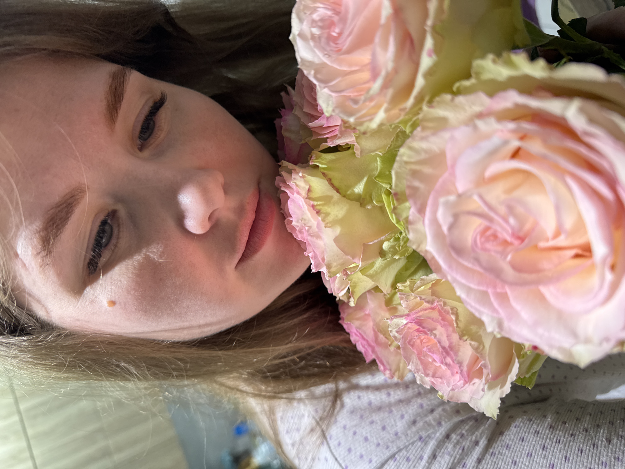 An image taken indoors in Gornyi (Dvurechenskoe S/P), Russia, depicts a close-up of a fair-skinned individual, likely female, with light brown hair. The subject's face is angled downwards, with eyes directed towards a bouquet of multiple light pink roses featuring greenish-white outer petals, positioned in the foreground. A distinct light brown mole is visible on the upper right side of the individual's face. The lips are lightly colored and slightly parted. The individual is wearing a light-colored garment with a subtle pattern of small, light purple dots on the left side of the frame. The background is out of focus, suggesting an interior setting with ambient lighting conditions.