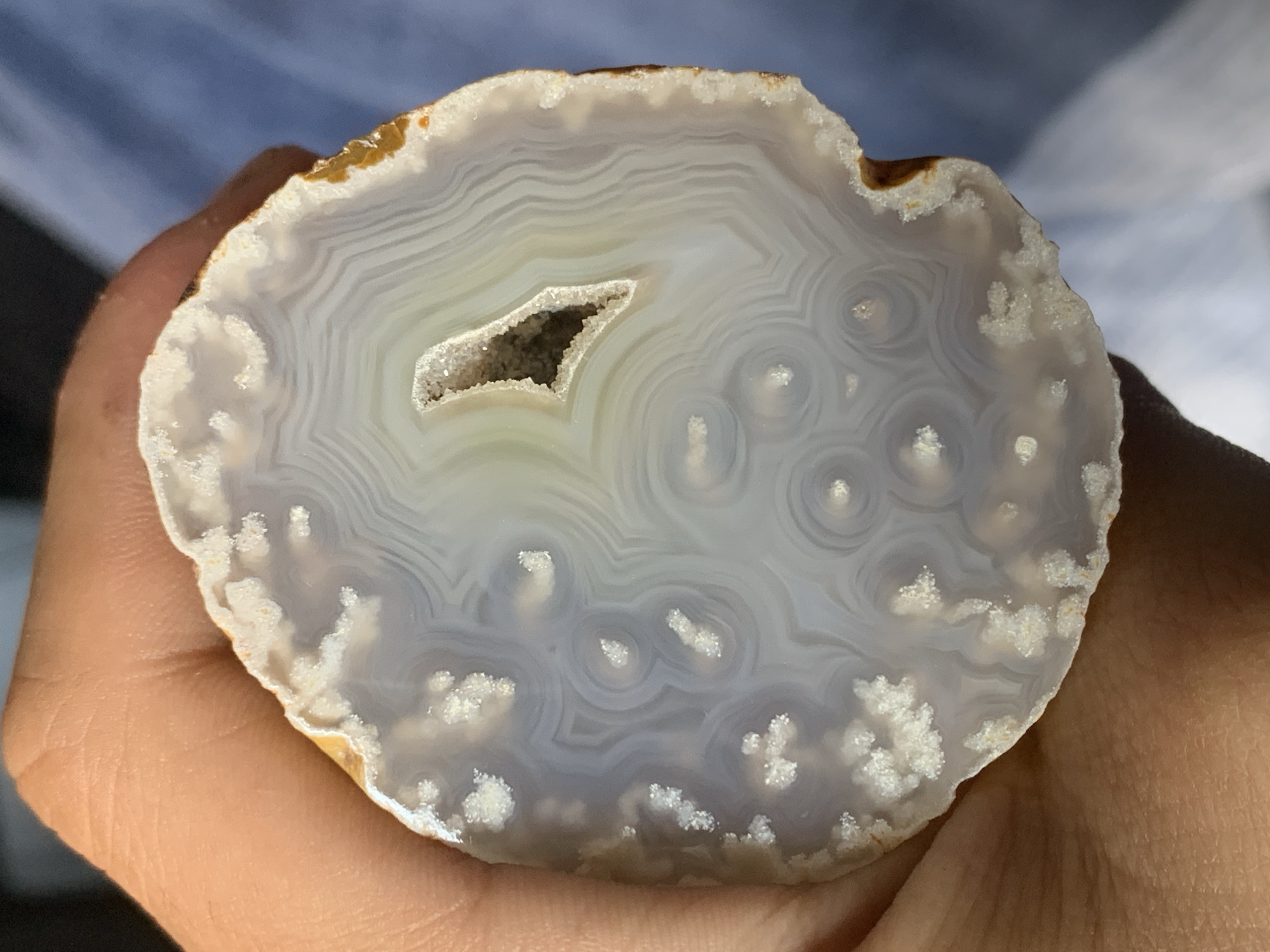 A hand holds a polished slice of an agate geode. The agate slice has a light gray and white banded pattern with a small druzy quartz-filled cavity in the center. The exterior edge of the agate is a brownish-orange color. The hand holding the agate has fair skin and visible wrinkles. The background is blurred, suggesting a light-colored surface, possibly fabric or sky. There is no indication in this image about Sofia, Bulgaria, or any specific location.