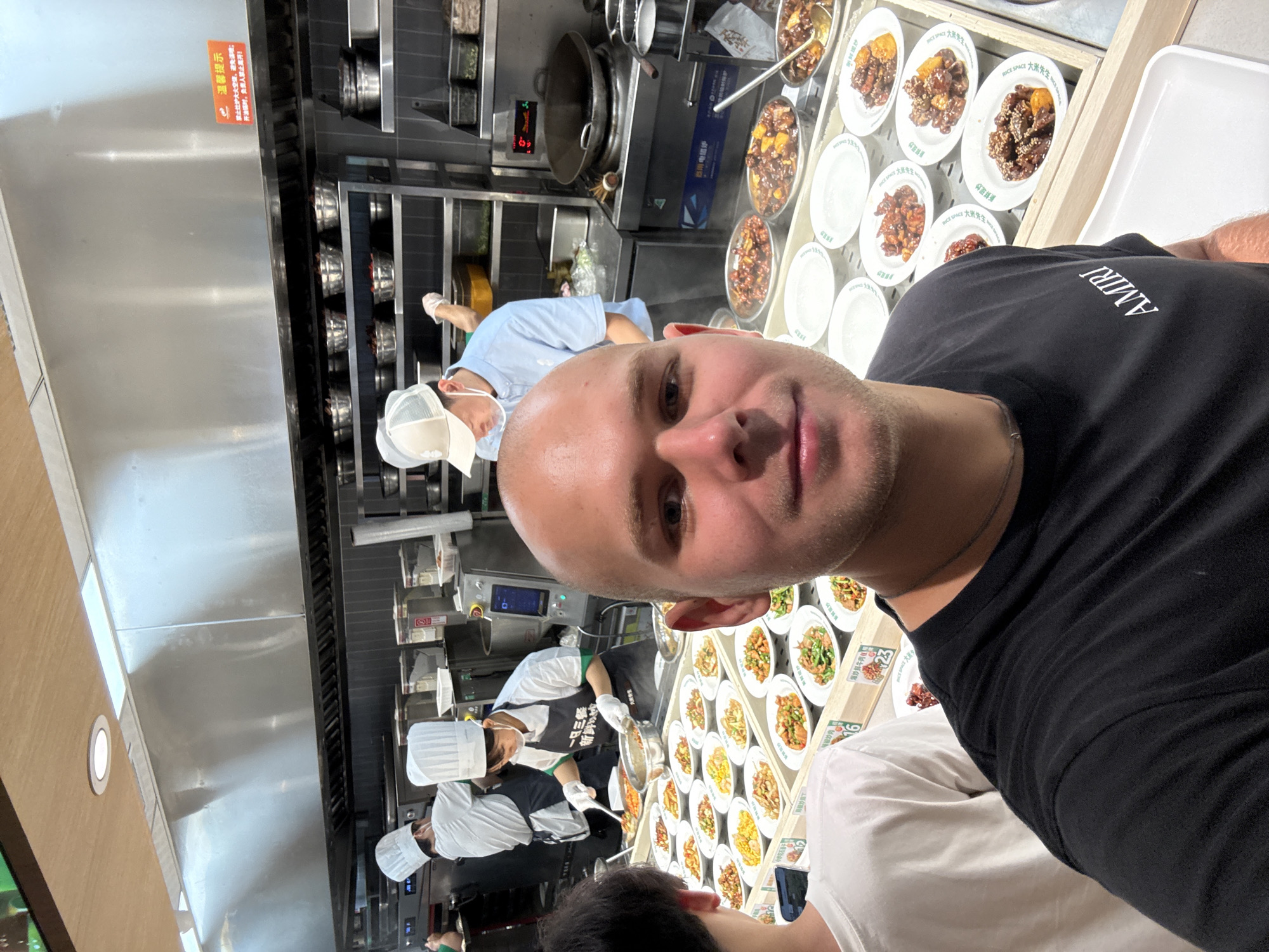 A bald, light-skinned man wearing a black t-shirt with the "AMIRI" logo visible on the left side of his chest, stands in the foreground and takes a selfie. He is in a brightly lit restaurant kitchen area where many pre-portioned dishes are displayed for selection. Behind him, three chefs in white hats and aprons are preparing and serving food. The kitchen features stainless steel counters, shelves stocked with ingredients and cookware, and a large exhaust hood. The man is looking directly at the camera. The scene appears to be in Tianhe District, China, suggested by the presence of Chinese signage and the style of the restaurant.