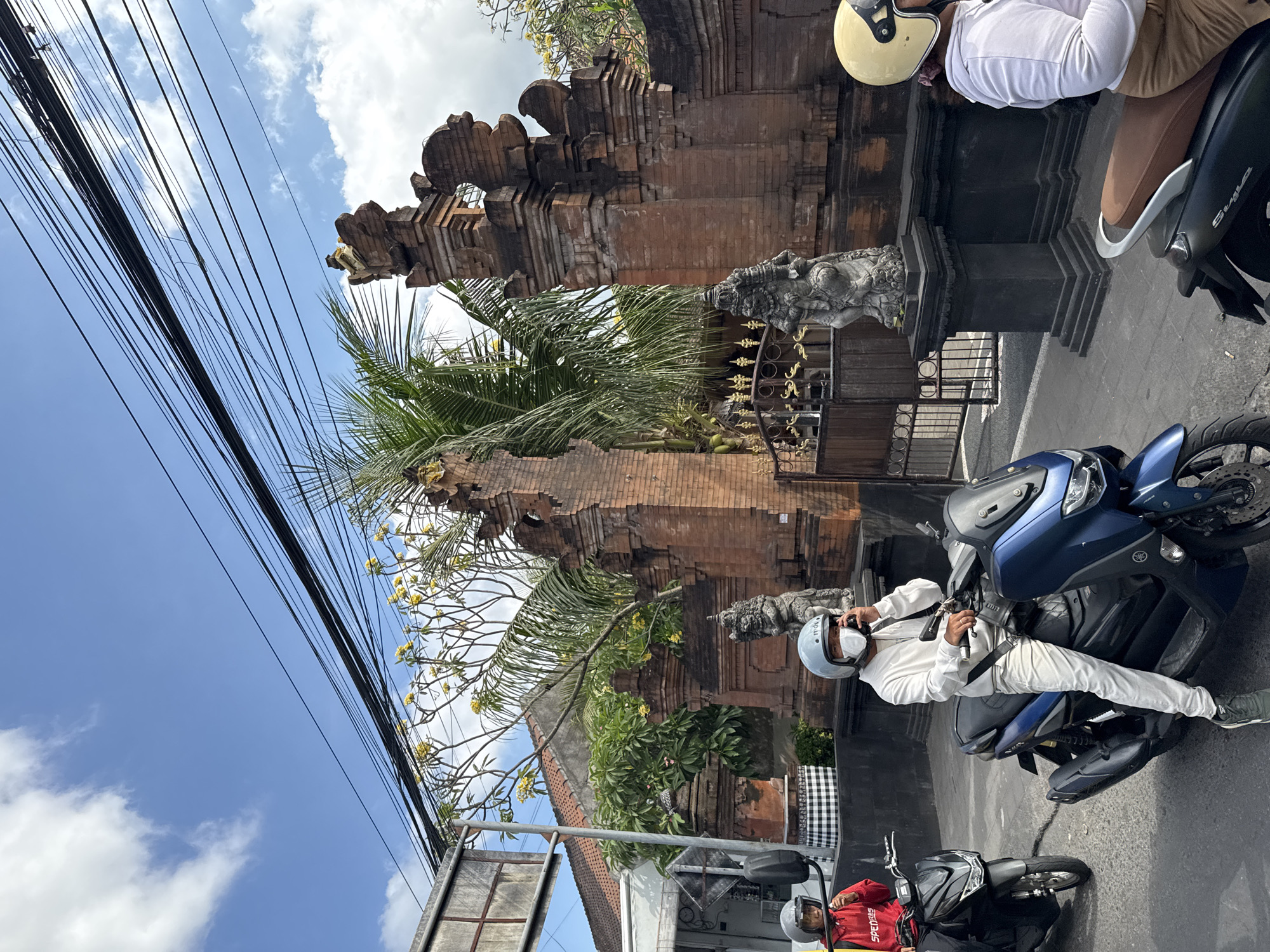 In Denpasar, Indonesia, under a bright blue sky with scattered clouds, a street scene unfolds featuring a group of people on motorbikes. A person in a red hooded sweatshirt with the word "SPORTS" visible is on a black motorbike to the left. In the center, a person dressed in a white hooded sweatshirt and white pants, wearing a light blue helmet and a white mask, is on a blue motorbike, appearing to be speaking on a mobile phone. To the right, another individual on a motorbike wears a light yellow helmet and a white hooded sweatshirt. The background is dominated by traditional Balinese brick gates, adorned with intricate carvings and sculptures, suggesting a cultural or historical setting. Lush green trees, some with yellow flowers, and palm fronds add to the tropical ambiance. Overhead, a dense network of electrical wires stretches across the sky, a common sight in urban Indonesian landscapes. The overall scene captures a moment of everyday life in Denpasar, with a blend of modern transportation and traditional architecture.