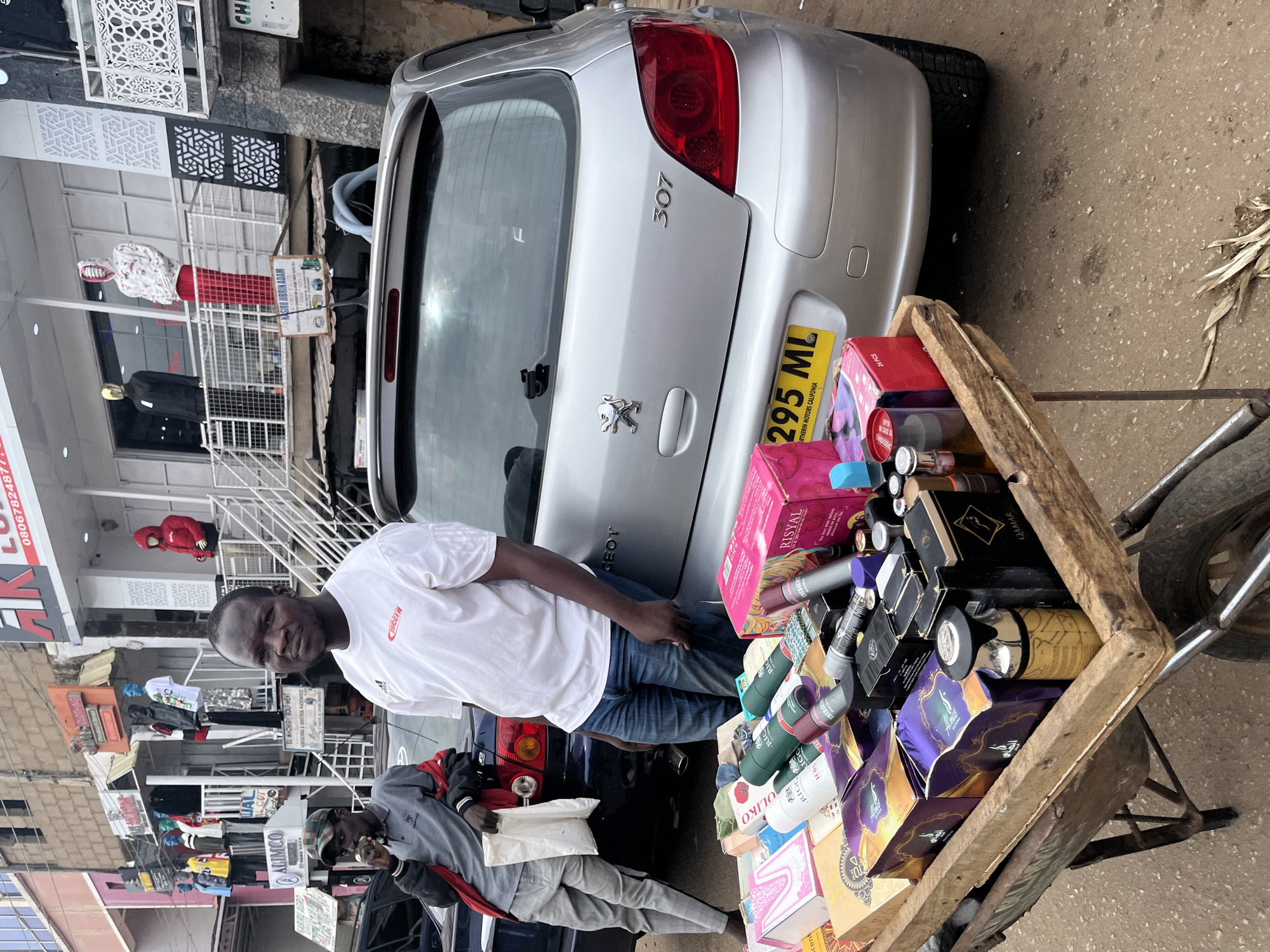 The image depicts an outdoor street scene in Jos, Nigeria, with a focus on a man selling various products from a cart parked in front of a silver Peugeot 307 hatchback. The man, wearing a white t-shirt with an Adidas logo and "SOCCER" text, and blue jeans, is standing to the right of the frame. To his left, another individual wearing a camouflage baseball cap, a gray jacket, and checkered trousers is leaning against a dark-colored vehicle.
The cart is laden with an assortment of products, including boxed perfumes, deodorants, and other cosmetics, some in purple and gold packaging. Prominent items on the cart include a large pink box labeled "RISYAL" and several cylindrical spray cans. The rear of the silver Peugeot is visible, displaying a license plate that reads "295 ML" and text that includes "NORTHERIN MOTORS CALIFORNIA."
In the background, buildings line the street, with shop fronts displaying various goods and mannequins dressed in clothing. The overall environment appears to be a busy commercial area, typical of a Nigerian urban setting. The ground is a mix of asphalt and dirt. The lighting suggests it is daytime, with some overcast conditions.