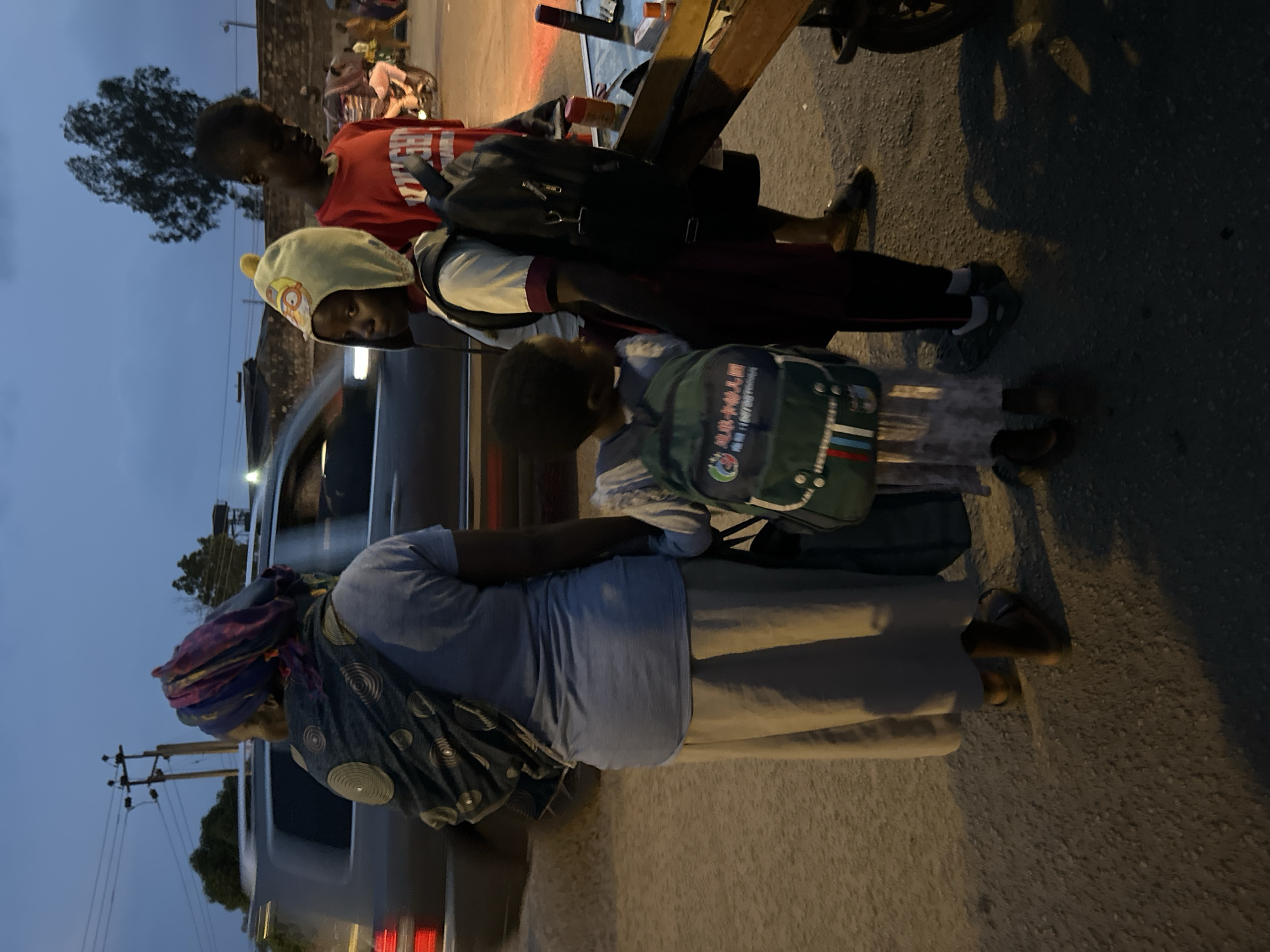 In the city of Bukuru, Nigeria, at dusk, an adult wearing a blue shirt and a light-colored skirt stands on a dirt road, facing away from the camera, with a patterned scarf draped over their head and shoulders. They are accompanied by two children, also facing away from the camera. One child, wearing a light-colored hat with cartoon characters, has a green backpack on their back, and the other child, wearing a red t-shirt, has a black backpack. Behind them, a car is partially visible. In the background, a tree and telephone poles are visible against a twilight sky. To the right, a wooden cart holds various items, including bottles and packaging. Other figures and a motorcycle are visible in the distance.