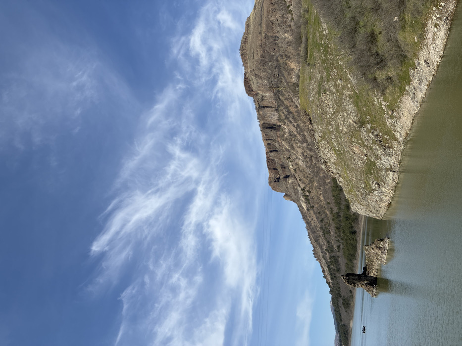 The image depicts a panoramic view of a mountainous reservoir landscape near Lisitsite, Bulgaria, under a partially cloudy blue sky. A calm body of greenish-brown water occupies the lower half of the frame. On the right, a steep, rocky bank covered with sparse green vegetation and bare shrubs slopes down to the water. Beyond this bank, a prominent mountain rises sharply, featuring a dark brown, vertically striated cliff face and slopes sparsely covered with dormant trees and patches of earth. In the mid-left section of the water, two jagged, dark brown rock formations protrude. Further to the left on the water, a small, dark-colored boat containing two discernible human figures is visible. Multiple thin, dark power lines traverse the middle-left sky horizontally. Distant hillsides with trees are visible on the far left, with faint suggestions of snow on higher peaks. The sky above is a bright blue, approximately 60% covered by white, wispy cirrus clouds.