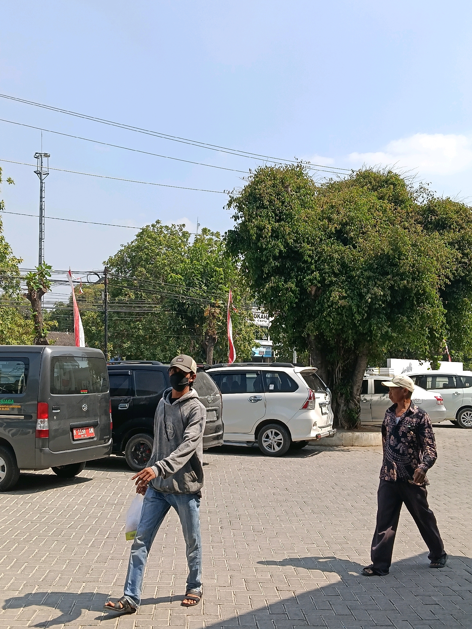 The image depicts an outdoor scene in Balun, Indonesia, characterized by a paved area with a brick pattern, likely a parking lot or public square. The day is clear with a bright blue sky, and sunlight casts distinct shadows on the ground.
In the foreground, two men are walking. The man on the left is younger, wearing a gray hooded sweatshirt, blue jeans, and black sandals. He has a dark face mask on and is carrying a translucent plastic bag in his right hand. He is facing towards the right side of the image. The man on the right is older, wearing a patterned dark shirt with a light-colored collar, dark pants, and dark sandals. He is wearing a light-colored baseball cap and is also facing towards the right.
Several vehicles are parked in the background. On the left, a dark gray van is visible, with a license plate that reads "K 1158 KM". Next to it is a black SUV. Further to the right, a white SUV is parked, followed by another white vehicle, possibly an SUV, on the far right.
The background features lush green trees with dense foliage, obscuring some of the structures behind them. Red and white flags, likely national flags, are displayed on poles, suggesting a public event or holiday. A tall communication tower or antenna stands to the left of the frame. The overall impression is a warm, sunny day in a public area with moderate vehicular traffic and pedestrian activity.
