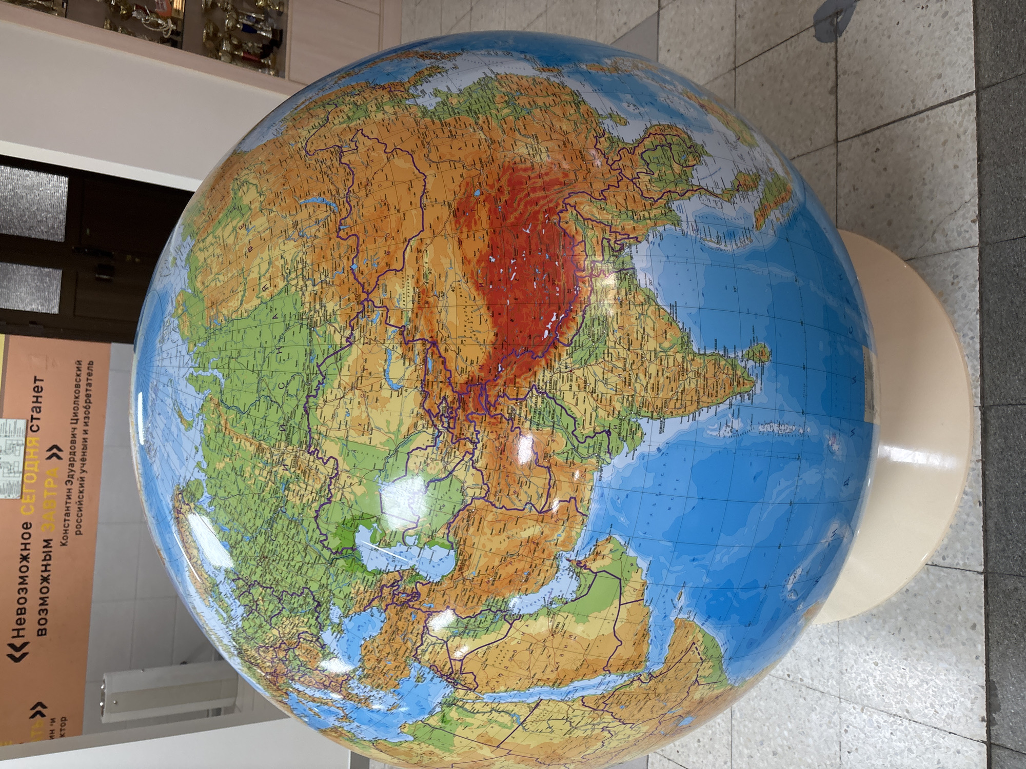 In Markovskii, Russia, a large, relief-style terrestrial globe, approximately 1.5 to 2 meters in diameter, occupies the foreground and center of the image. The globe displays a physical map of Earth, predominantly featuring Eurasia, Africa, and parts of Oceania. Oceans are depicted in blue, while landmasses show varying elevations with shades of green, yellow, brown, and a prominent red area highlighting the Himalayas. Political borders, major cities, rivers, and geographical names in Cyrillic script are visible across the surface. The globe rests on a cream-colored, circular pedestal base.
The background consists of a tiled floor with light gray and darker gray square tiles. Against the left wall, an orange-beige panel displays Cyrillic text in black, reading: "<<Невозможное СЕГОДНЯ станет возможным ЗАВТРА >> Константин Эдуардович Циолковский российский ученый и изобретатель" (translating to: "<<The impossible TODAY will become possible TOMORROW >> Konstantin Eduardovich Tsiolkovsky Russian scientist and inventor"). To the right, a dark-colored door with frosted glass panels is partially visible, adjacent to a white wall corner. Further right, a glass display cabinet containing multiple gold-colored trophies or awards is visible. No individuals are present, and no actions or interactions are depicted within the scene.