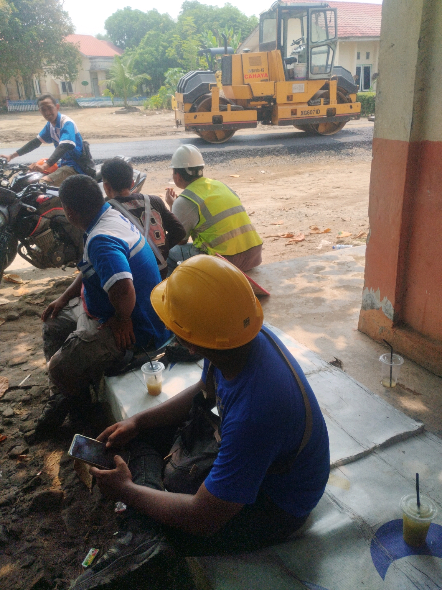 In Pagarbosi, Indonesia, a group of construction workers are gathered near a road construction site. In the foreground, a man wearing a yellow hard hat and a blue shirt sits on a makeshift bench, looking at his phone. Beside him, another man in a blue and white shirt and khaki pants is also seated, and a third man in a black shirt with a red number 8 on the back is visible behind him. In the background, a yellow steamroller is operating on a freshly paved road. Another worker in a bright yellow vest is standing near the steamroller. The scene is outdoors, with a building and trees visible in the background, indicating it's a sunny day. The ground is a mix of dirt and paved surfaces, with debris scattered around.