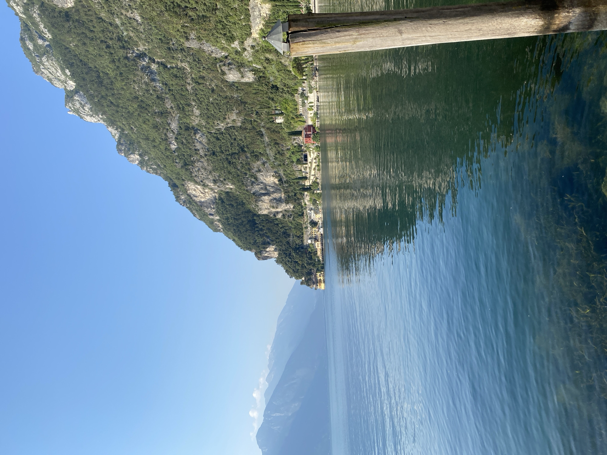 This is a medium shot photograph taken from a body of water, likely Lake Garda, looking towards the shore of Riva del Garda, Italy. In the foreground, a weathered wooden post with a dark, octagonal metal cap juts out of the water on the right side of the frame. The water itself is a deep blue-green, with the clear bottom visible in some areas, revealing submerged aquatic plants. Ripples on the surface create reflections of the surrounding landscape.
The middle ground features a picturesque shoreline with buildings nestled at the base of a steep, lush green mountainside. The buildings appear to be a mix of residential and possibly commercial structures, with some showcasing colorful facades, including a prominent red building. Terraced gardens and cypress trees dot the landscape, climbing the slopes.
The background is dominated by the imposing, rocky mountain range that forms the backdrop to the town. The slopes are densely covered in a mix of trees and exposed rock faces, with visible paths or roads winding their way up. The sky is a clear, bright blue with a few faint wisps of clouds in the distance. The overall impression is one of a serene, sunlit day in a beautiful natural setting.